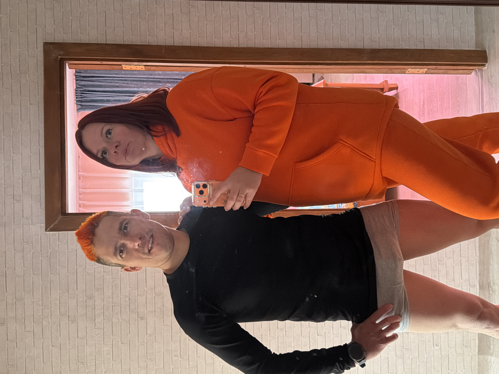 Two people pose for a selfie in front of a mirror. A woman with shoulder-length, dark red hair wears an orange hoodie and holds up a cell phone with an orange case. Below her, a man with short, orange hair and a black long-sleeved shirt stands with his hand on his hip. He wears light gray shorts. The reflection in the mirror shows curtains and a pinkish hue. The background consists of a white brick-patterned wall, and a wooden-framed doorway. There is no verifiable information to indicate the location is Chaikovsky, Russia.