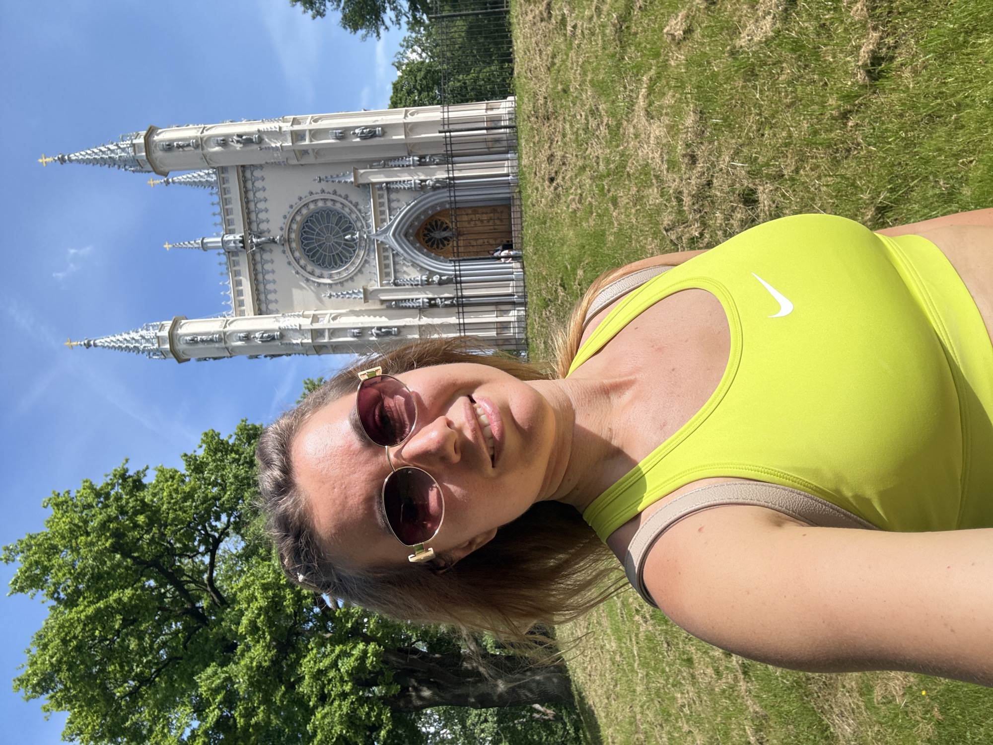 An outdoor eye-level selfie captures a female subject in the foreground with the Gothic Chapel in Alexandria Park, Peterhof, St. Petersburg, Russia, in the background on a sunny day.
The subject, an adult female with long light brown hair, wears large, oval-shaped sunglasses with gold frames and dark lenses, smiling directly at the camera. She is clad in a bright lime green sports bra or top featuring a white Nike swoosh logo on the front. A light-colored strap, possibly from a backpack, is visible over her right shoulder. Her skin shows a tanned complexion.
The background prominently features the Neo-Gothic style Gothic Chapel. The chapel, constructed from light-colored material, exhibits multiple ornate spires topped with golden Orthodox crosses, a large circular rose window with intricate tracery above the main entrance, and a pointed arched wooden doorway. Numerous statues are visible within niches along its facade. A dark metal fence encloses the chapel at its base, with two small, distant figures visible near the entrance.
To the left, a large, mature tree with dense green foliage occupies the mid-ground. The foreground and mid-ground consist of a grassy slope, with visible patches of cut grass. The sky is clear blue with a few wispy white clouds. The scene is brightly illuminated by natural sunlight.