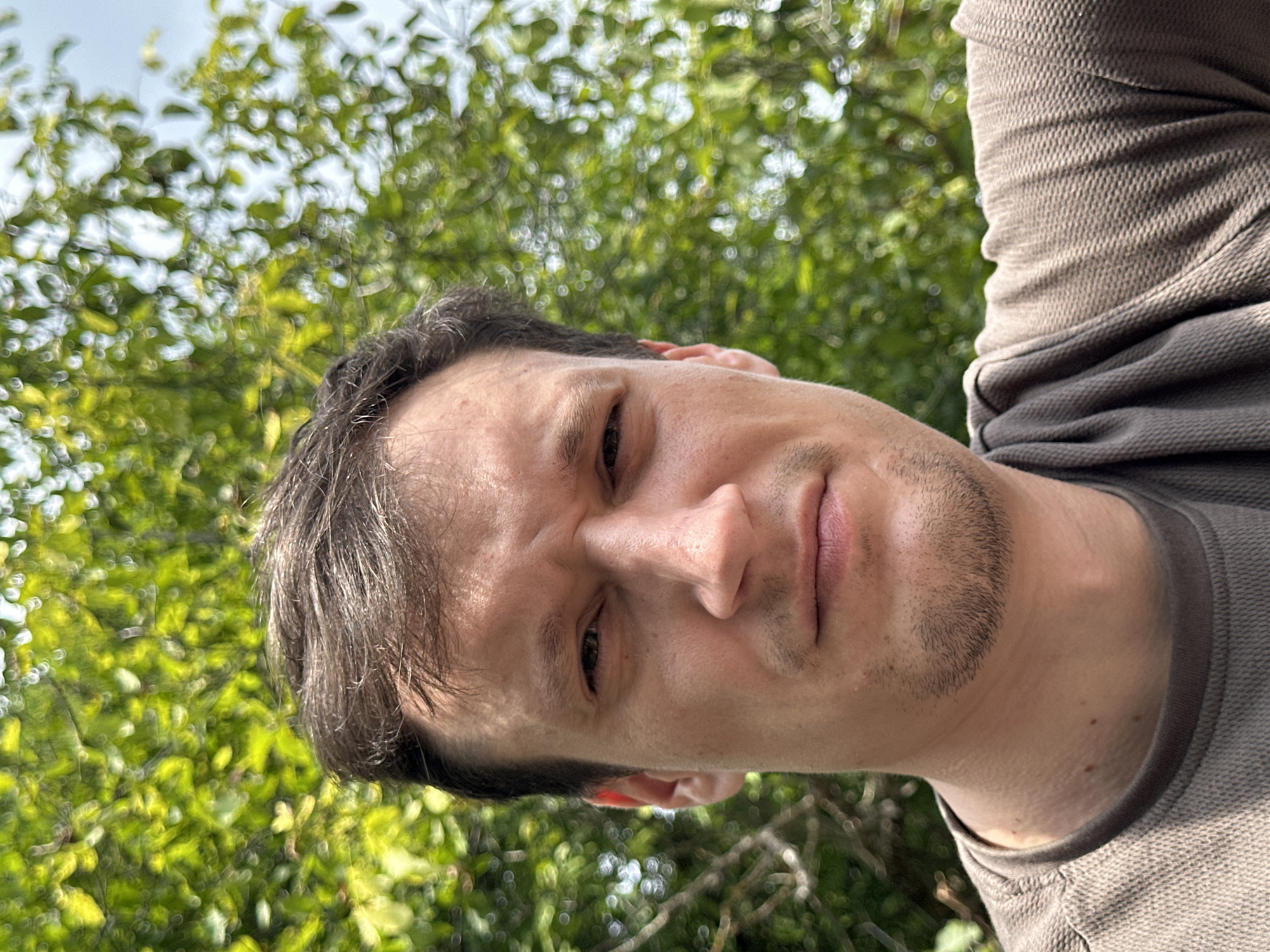 In a close-up, self-taken photograph against a backdrop of blurred green foliage and a hint of sky, a man with short, brown hair and a light stubble appears. He is wearing a brown, textured crew-neck shirt. His facial expression is neutral, with his eyes looking directly at the camera. The lighting suggests an outdoor setting, possibly during the daytime. The exact location within the city of Makiv, Ukraine, is not discernible from the image alone.