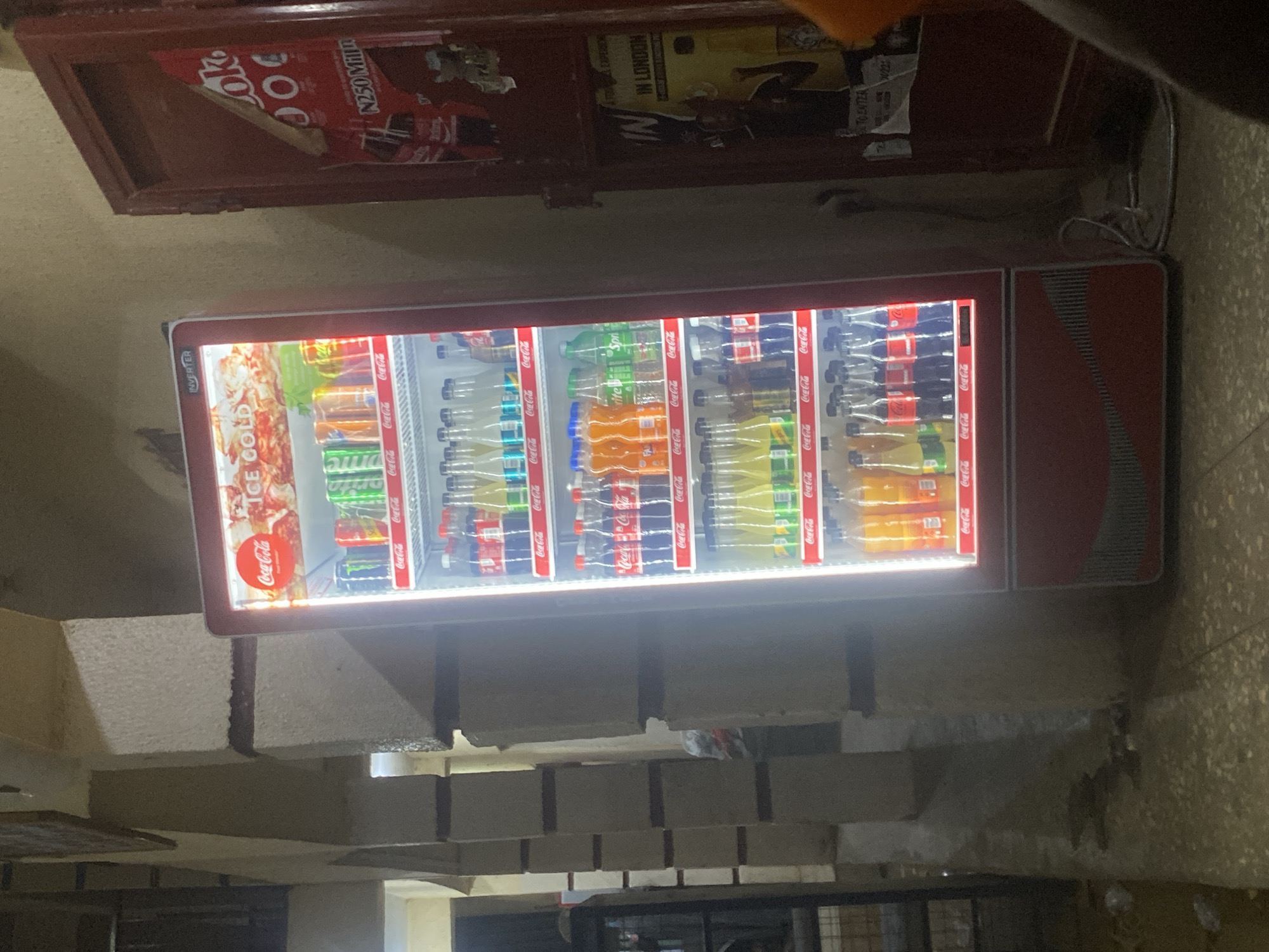 Here is a forensic description of the image.
**Location:** The image appears to be taken in Tukur Tukur, Nigeria, based on information from the Coca-Cola cooler and a 'N250 Milln' sign which likely refers to Nigerian currency and products.
**Subjects and Objects:**
* A Coca-Cola refrigerator or cooler. The refrigerator is stocked with Coca-Cola products, including various flavors of Coca-Cola and Sprite. There are some food products visible at the top section of the cooler.
* Building structure: The background shows a portion of the building structure with a set of steps leading somewhere.
**Description:**
The primary focus of the image is a Coca-Cola refrigerator positioned against a textured wall. The refrigerator is well-lit, displaying a range of beverage products. Above the refrigerator is a shelf with Coca-Cola marketing and another poster mentioning 'IN LONDON'
The refrigerator is sitting in front of some steps or some building supports of some sort. The scene suggests the photo was taken in an urban setting in Nigeria, likely a local store or distribution point for Coca-Cola products.