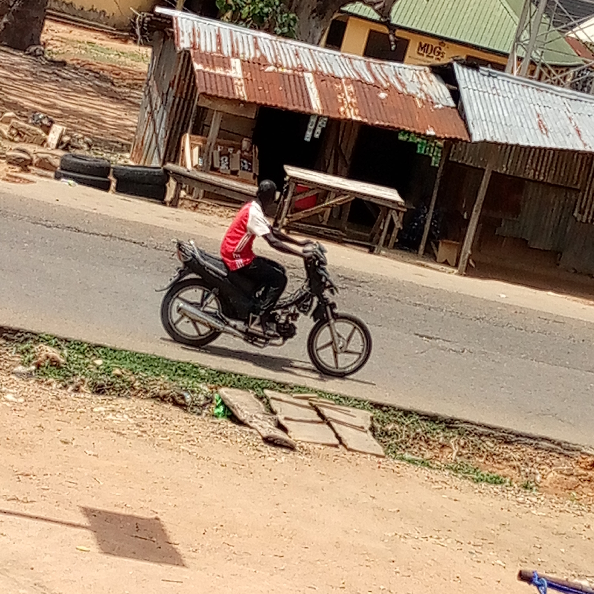 The image shows a man riding a black motorcycle on an asphalt road. He wears a red shirt with white accents and dark trousers. The road is flanked by grassy areas and a dirt surface. In the background, there are several makeshift shops with corrugated iron roofs. One of the shops has the letters "MDGs" visible on its facade. There are also a couple of tires on the side of the road near one of the shops. The location is identified as an urban area in the city of Unknown, Unknown.