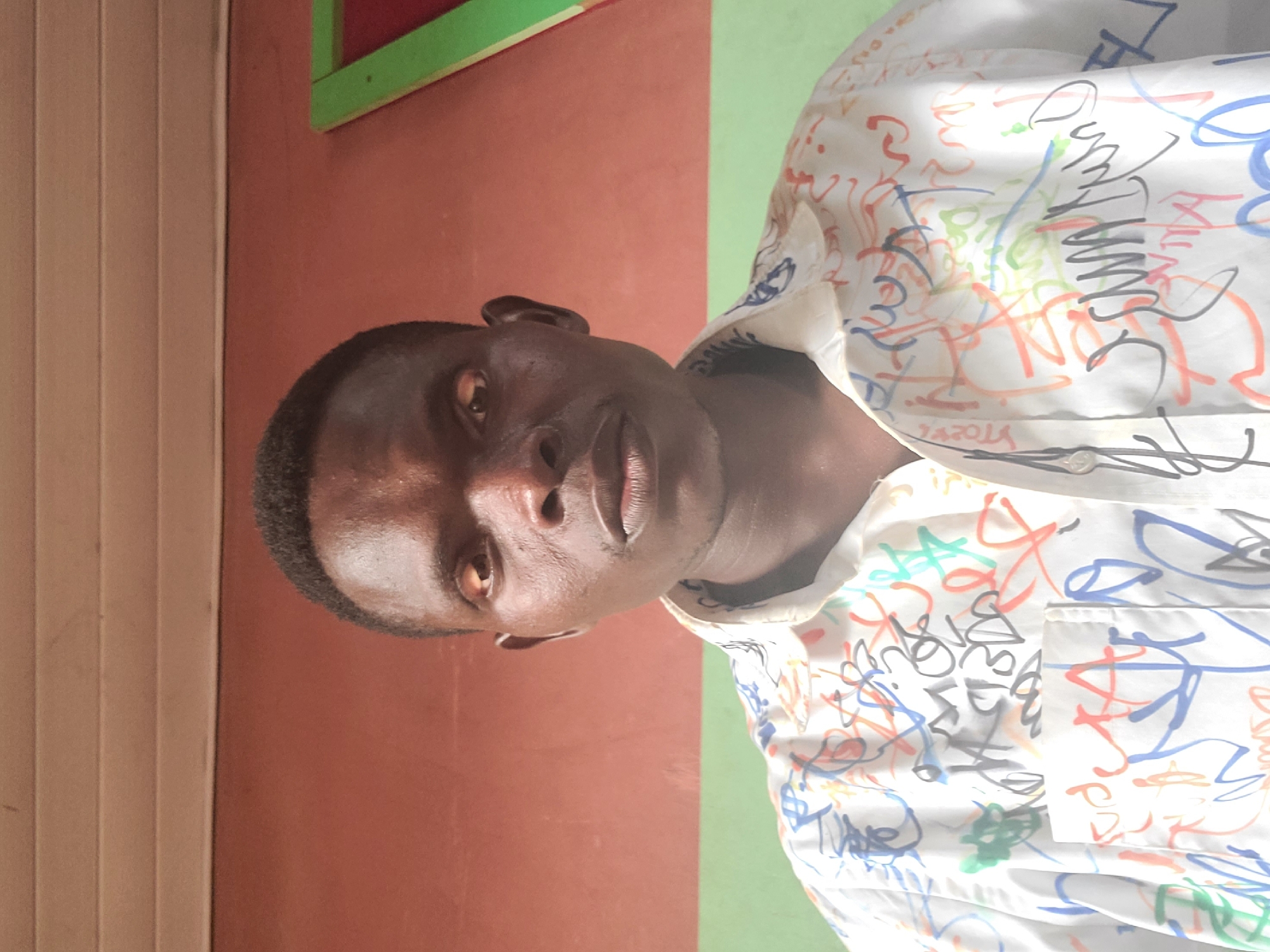 A medium-shot frontal portrait of a Black man in his late 20s or early 30s is captured in Gura Top, Nigeria. He has short, dark hair, brown eyes, and dark skin. He is wearing a white button-down shirt with a colorful graffiti-style pattern of red, blue, green, and orange squiggles and shapes. The background consists of a wall with a horizontal wood paneling at the top, painted a light tan color. Below this, the wall is painted a dusty rose color and then a muted green, separated by a horizontal line. To the right of the man's head, a portion of a green-framed window with a red interior is visible. The lighting suggests an indoor setting with natural light coming from the front.