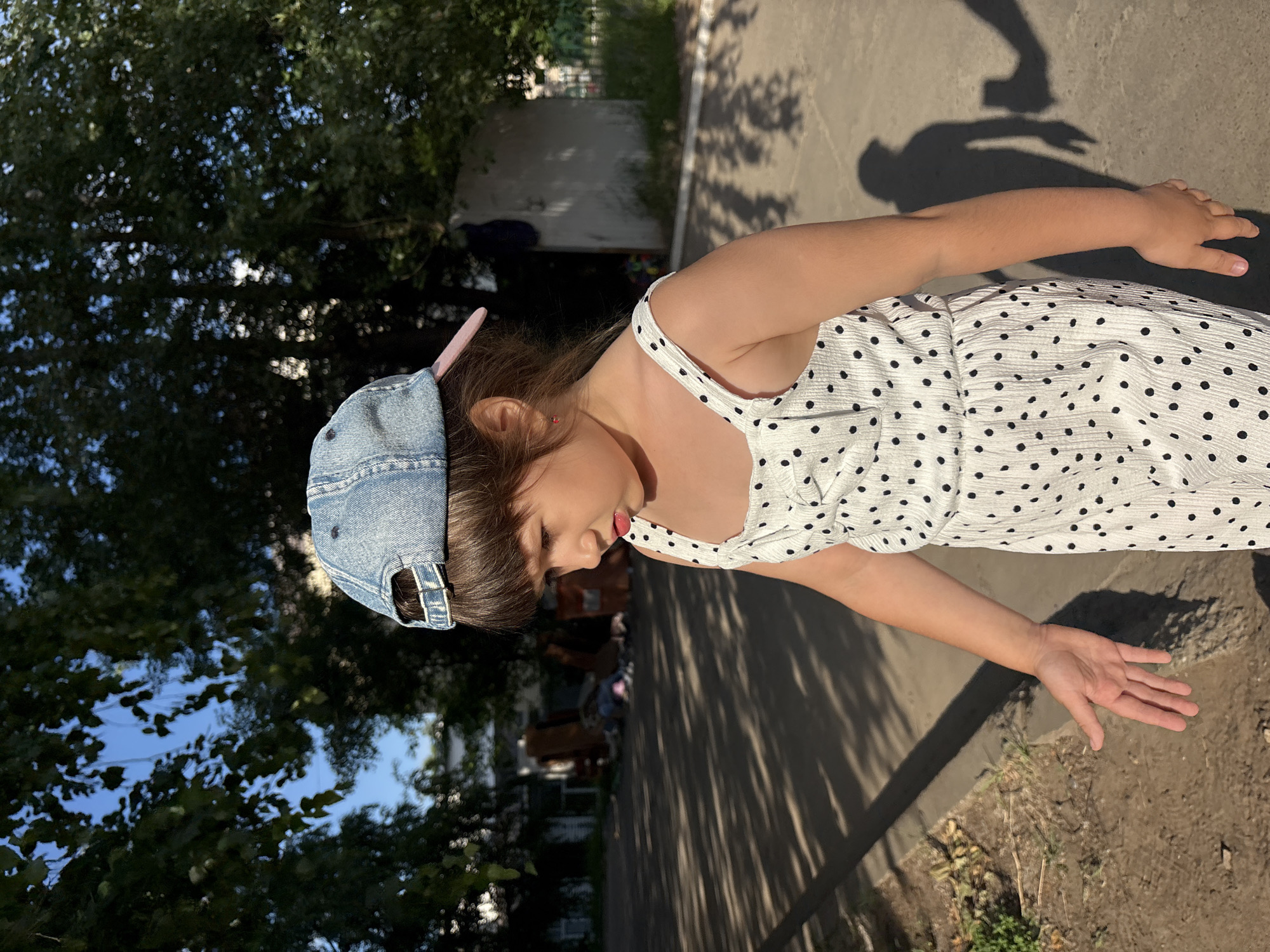 A young girl wearing a denim baseball cap and a white polka-dotted dress is standing outdoors on a paved surface. She is looking down and to her right. The background shows trees, buildings, and debris. The lighting suggests it is daytime, with shadows cast on the ground. The location is identified as Chaikovsky, Russia.