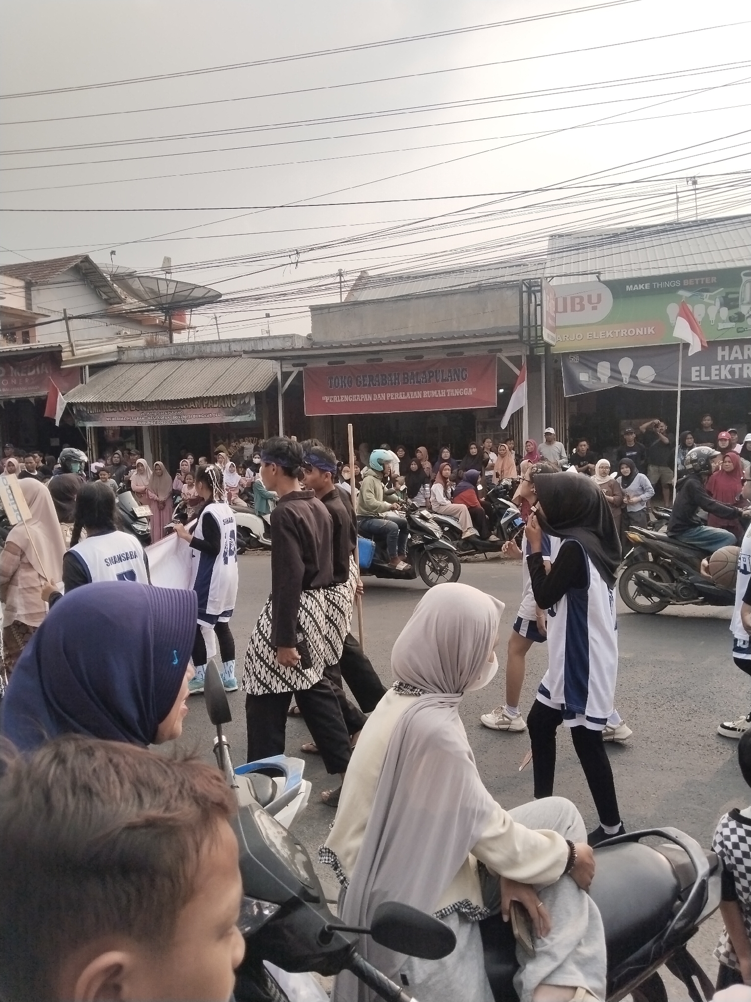 The image depicts a street scene in Balapulang Kulon, Indonesia, likely during a procession or festival. A crowd of people, many wearing hijabs and traditional Indonesian clothing, are gathered along a street lined with shops. Several individuals are walking or standing, while others are on motorcycles, some of which are adorned with decorations. In the foreground, two children are visible, one on a motorcycle. The shops appear to be a mix of retail stores, including one with a banner advertising electronics. The sky is overcast, and power lines are strung across the top of the frame. The overall atmosphere suggests a lively community event.