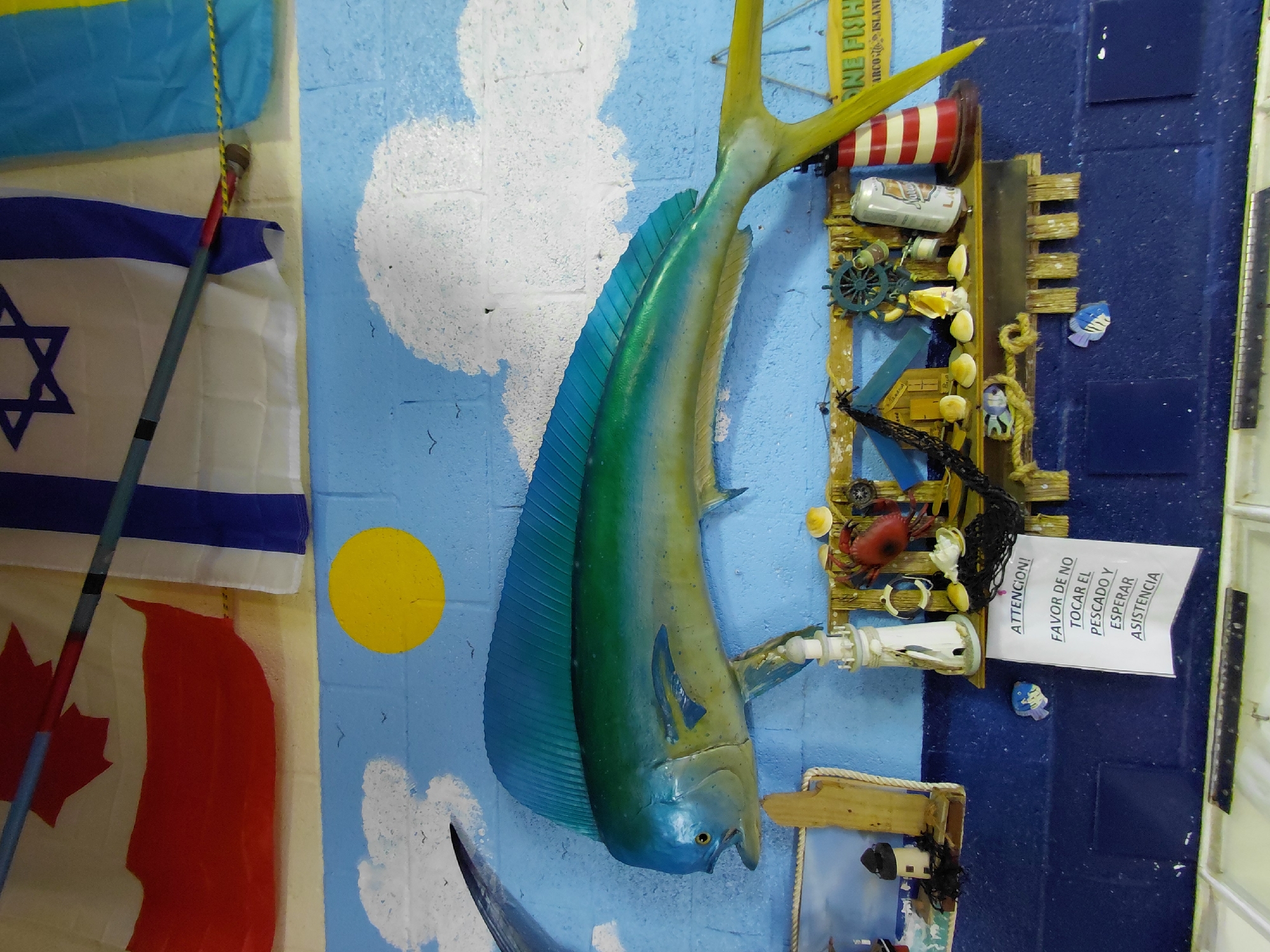 The image shows a wall display with a nautical theme. A large, brightly colored mahi-mahi (dolphin fish) replica dominates the center, mounted against a painted backdrop of light blue sky with white clouds and a yellow sun. To the left of the fish are flags of Canada, Israel, and Ukraine, hanging vertically. To the right of the fish is a wooden structure decorated with nautical items like a miniature lighthouse, a crab, shells, and a beer can, along with a sign in Spanish reading, "ATTENCION! FAVOR DE NO TOCAR EL PESCADO Y ESPERAR ASISTENCIA" ("Attention! Please do not touch the fish and wait for assistance"). Above the fish is a sign that says “ONE FISH MARCO ISLAND”. Based on context clues, the image was likely taken in Cutler Bay, United States of America.