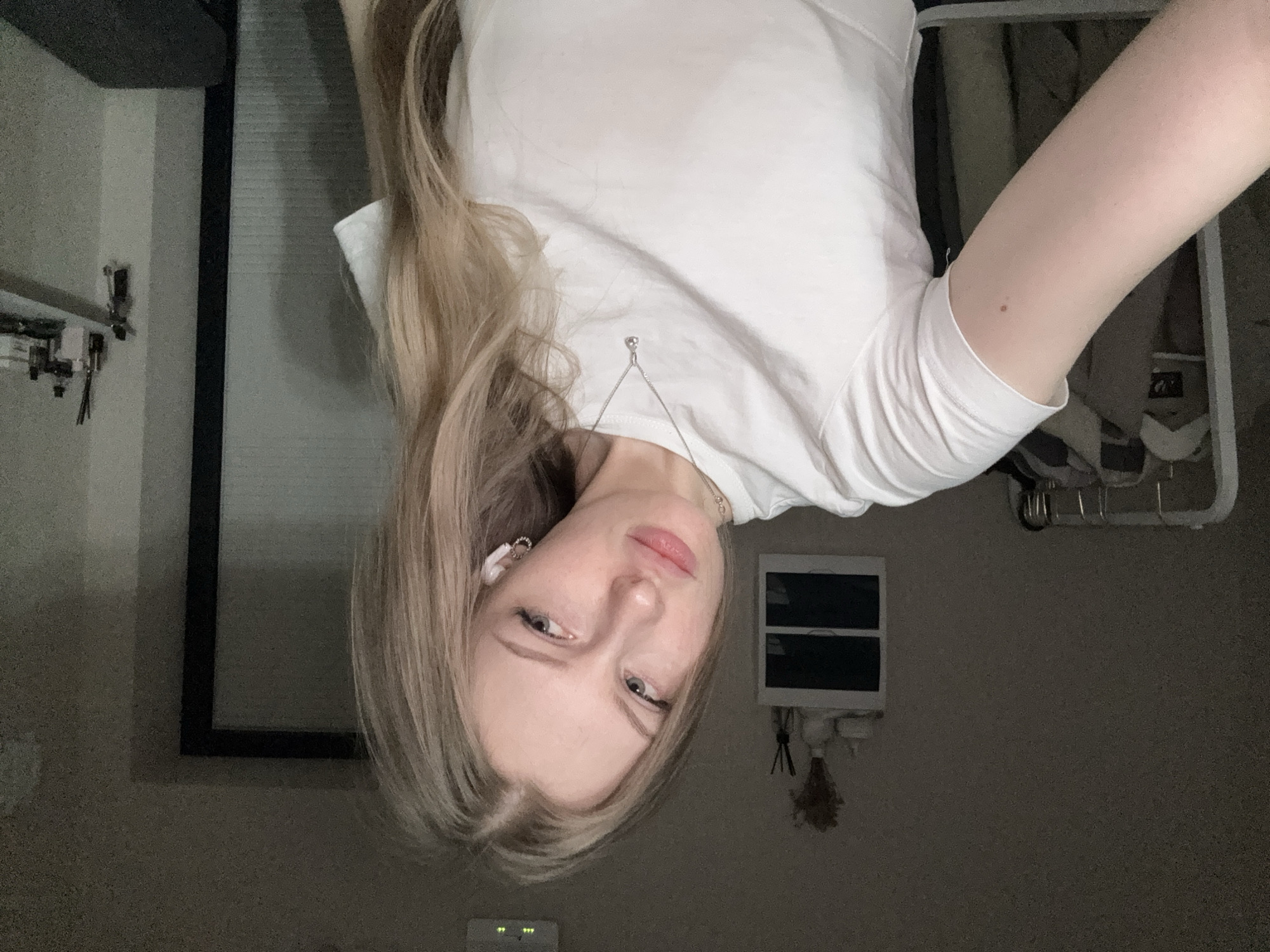 The image features a young woman with long, straight blonde hair and fair skin, looking directly at the viewer. She is wearing a white short-sleeved t-shirt and a delicate silver necklace with a small, round pendant. A white earbud is visible in her left ear, suggesting she might be listening to something. Her expression is neutral to slightly contemplative, with her lips subtly parted.
The setting is an indoor environment, likely a room in a home. The lighting is dim and appears to be artificial, implying it may be evening or a space with limited natural light. In the background, to the woman's left, there is a light-colored wall with what appears to be a dark-framed window or mirror. To her right, vaguely discernible elements suggest a closet or clothes rack with hangers. Below this, a small, dark decorative item, possibly dried flowers, hangs on the wall, alongside a white rectangular object. In the lower-central part of the background, a digital display is visible, showing "5:00" and three small dots beneath it, which could indicate the time. The overall atmosphere is casual and personal, consistent with a self-portrait taken in a private space. The location context provided is Perm, Russia.