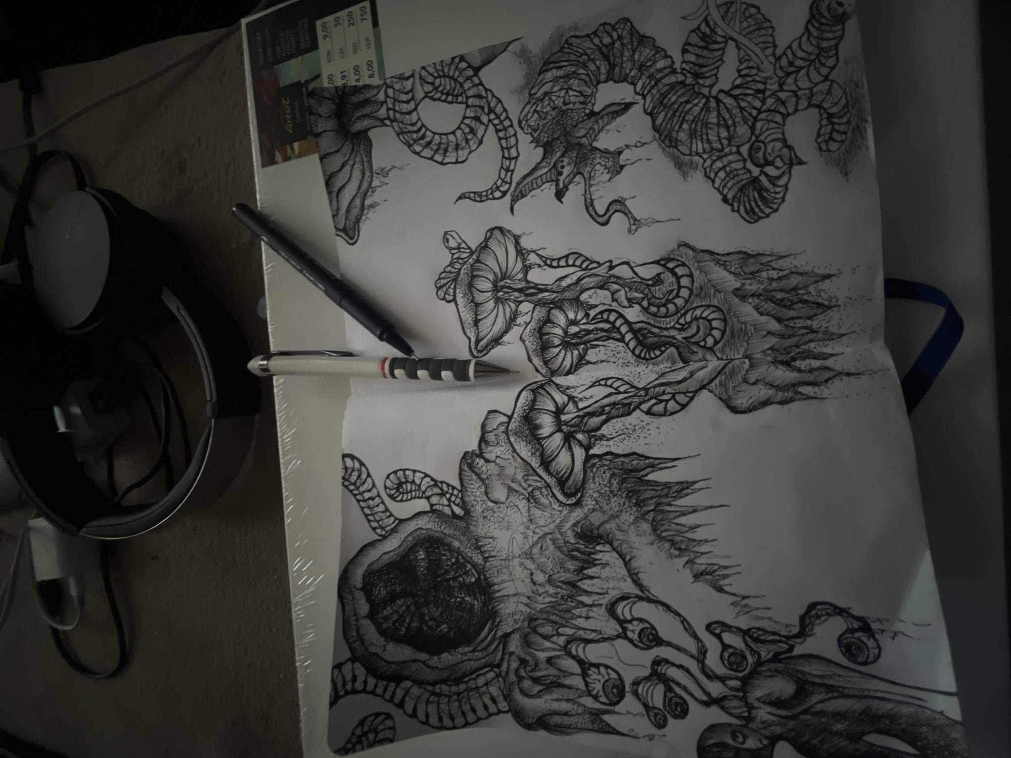 This indoor image presents a detailed black and white fantasy drawing laid flat on a surface, likely a desk or table, indicating an artistic activity. The intricate artwork features organic, fantastical forms, resembling monstrous creatures with tentacles, eyes, and textured elements, rendered using fine lines, cross-hatching, and stippling. Two pens, one black and one white with black stripes, rest on the drawing.
To the left, a pair of black over-ear headphones with a silver/grey ear cup, a white charging block, and cables are visible, suggesting a personal workspace. Behind the drawing, a sheet of paper is partially obscured, displaying what appears to be a price list with various currency codes and numbers. Visible text includes "Artist," "CMRVAS," "PANE," "SMOOTH," and currency information such as "RON 9,00," "3.91 CZK 50," "4,00 RSD 250," and "8,00 HUF 750," which provides regional location cues consistent with Sofia, Bulgaria. The image is dimly lit, casting soft shadows and contributing to a cool, muted atmosphere. A blue strap or handle is visible on the far right.