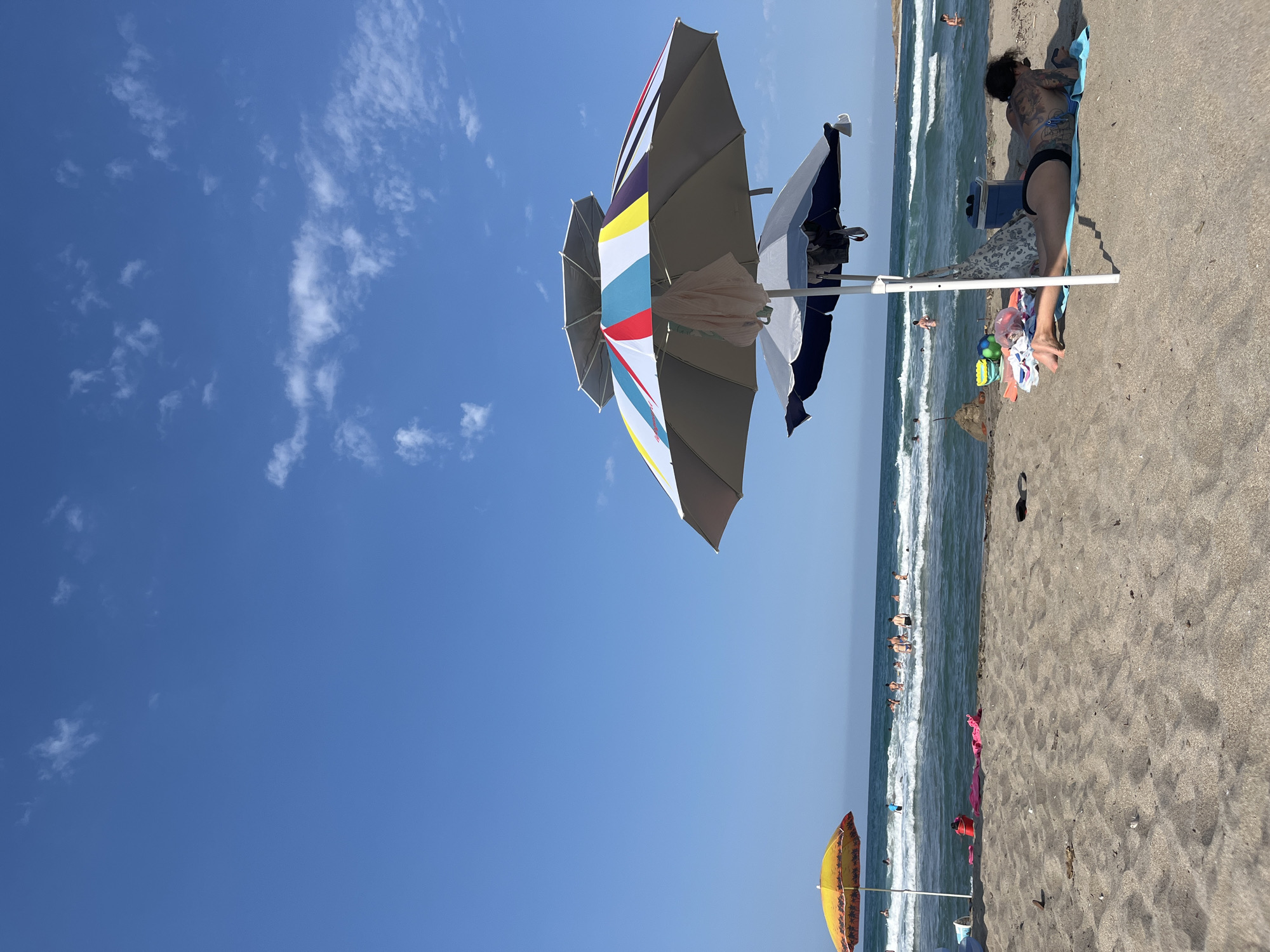 The image depicts a sunny day at Ahtopol, Bulgaria, with a clear blue sky and scattered white clouds. The scene is a beach with people enjoying the water and sand.
The main subjects are the beachgoers and beach accessories. In the foreground, several beach umbrellas are visible, including one with a colorful striped pattern and another in blue and white. There are also people relaxing on towels on the sand. One person, with numerous tattoos and wearing black bikini bottoms, is lying on a blue towel, facing away from the camera. Near them, beach bags, a cooler, and various colorful items are scattered on the sand.
In the background, the sea is visible with moderate waves and a clear horizon. Several people are in the water, either swimming or wading. A yellow umbrella is also present on the beach.
The overall atmosphere is one of leisure and relaxation on a warm day. The bright sunlight suggests it is midday. The presence of the sea and umbrellas confirms the beach setting. No visible text is present in the image.