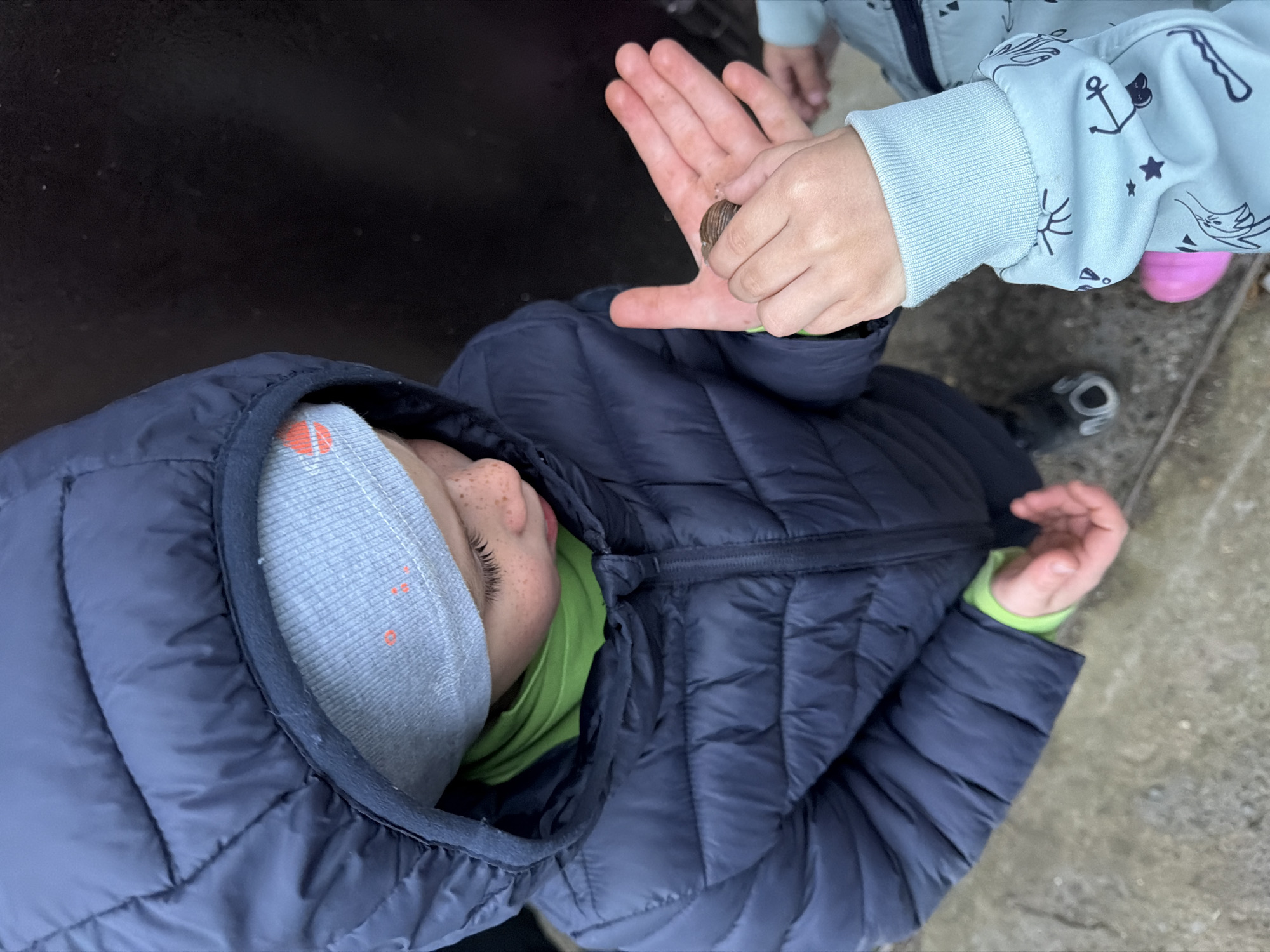 A high-angle shot depicts two children, one holding a snail in their outstretched hand. The child closest to the camera is wearing a navy blue puffer jacket with a gray beanie and a lime green shirt underneath. The second child, partially visible, wears a light blue jacket adorned with marine-themed patterns. A small, brown snail sits on the open palm of the second child's hand. The setting appears to be outdoors, with a dark, possibly metallic surface on the left and a gray concrete area on the right.