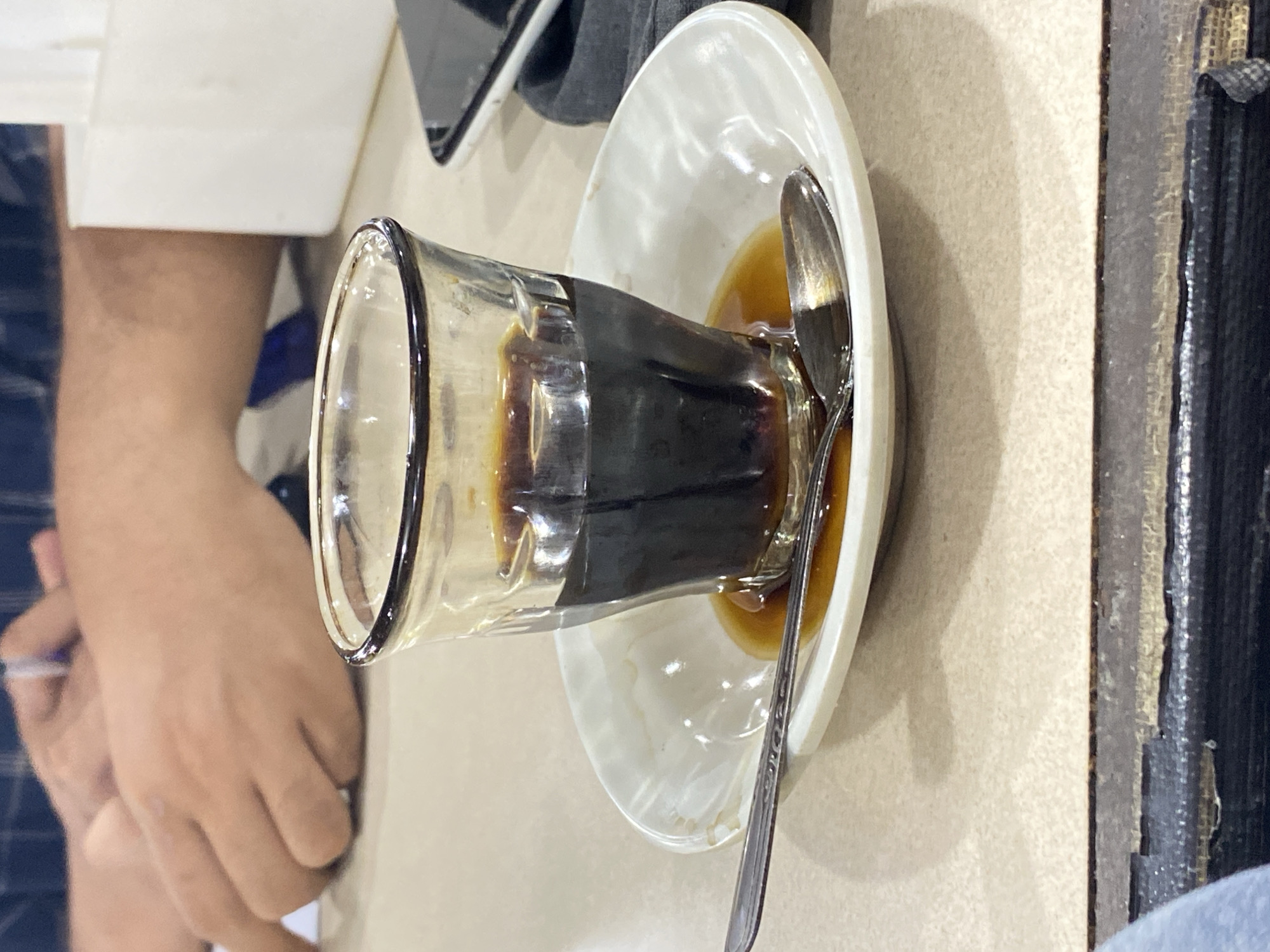The image captures a close-up view of a small glass of dark coffee placed on a white saucer. A silver spoon rests on the saucer, partially submerged in a small pool of coffee that has spilled around the base of the glass. The coffee in the glass is dark and opaque, with some reflections visible on its surface. The saucer has a slight sheen and appears to be ceramic. In the background, out of focus, are parts of a person's hands and arms, suggesting someone is seated at the table. The person is wearing a dark, patterned shirt. To the right, a white object, possibly a tissue box or a dispenser, and a portion of a smartphone are also visible. The surface of the table is a light-colored, textured material. The overall lighting suggests an indoor setting, possibly a cafe or restaurant. The scene is set on a table in a cafe in Lhokseumawe, Indonesia.
