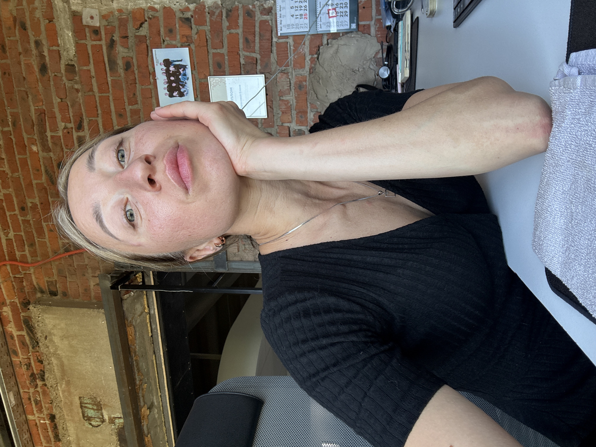 The image shows a woman in a black top, seated at a desk, resting her chin on her hand. She has blonde hair and is looking towards the camera. The background consists of a brick wall with a calendar and a framed photo of a group of people. There is also a chair with a grey fabric seat and headrest. The scene appears to be an indoor workspace. There is no identifiable information about the location in the city of Chaikovsky, Russia.