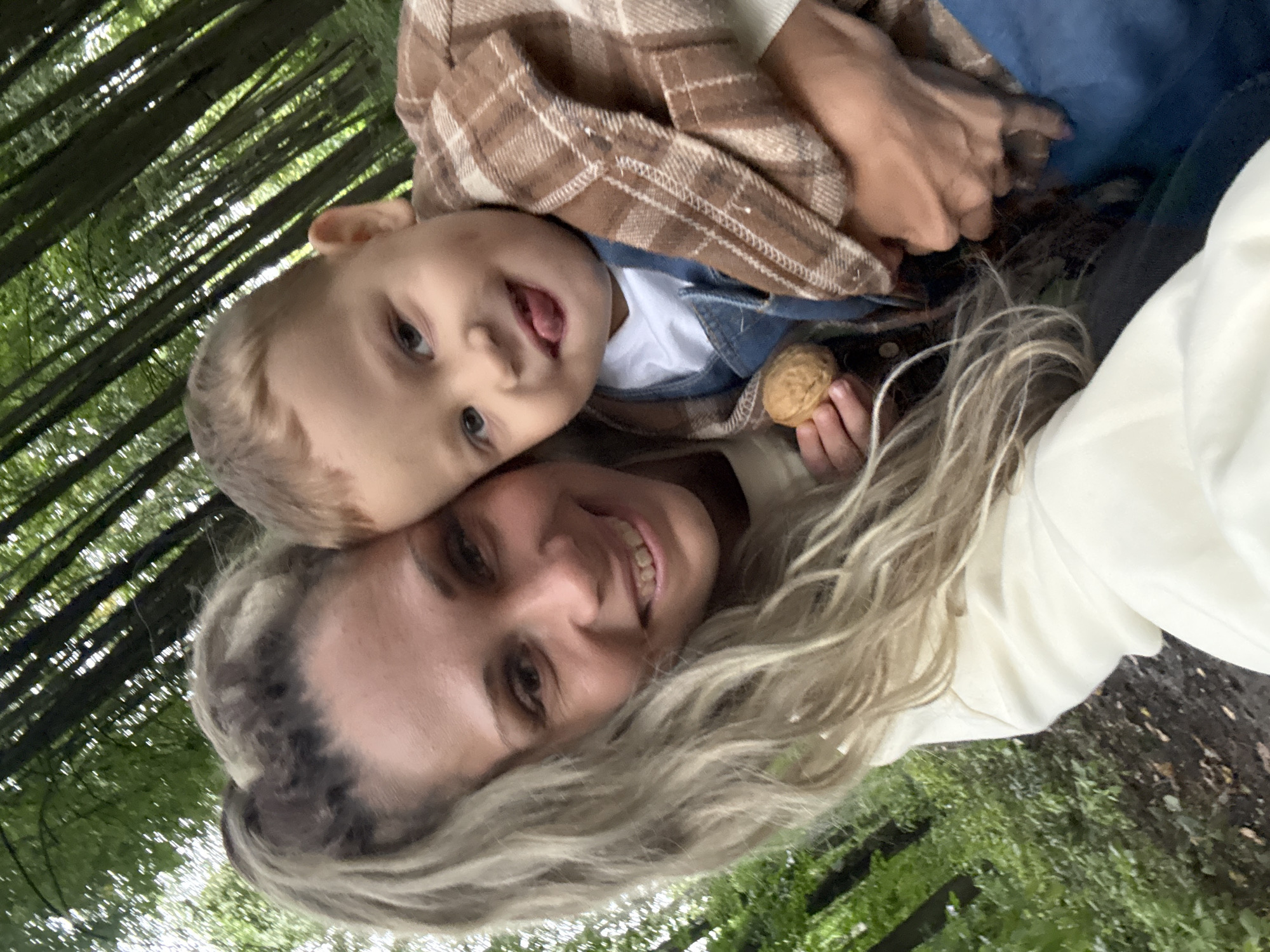 A selfie is taken by a woman with blonde, wavy hair tied in a ponytail, who is smiling. She is holding a young child who is sticking out his tongue. The child is wearing a plaid shirt and denim overalls. In the child's hand is a walnut. They are in a forest with tall trees and green foliage, likely in or near Rusanovo, Russia. The ground appears to be a dirt path.