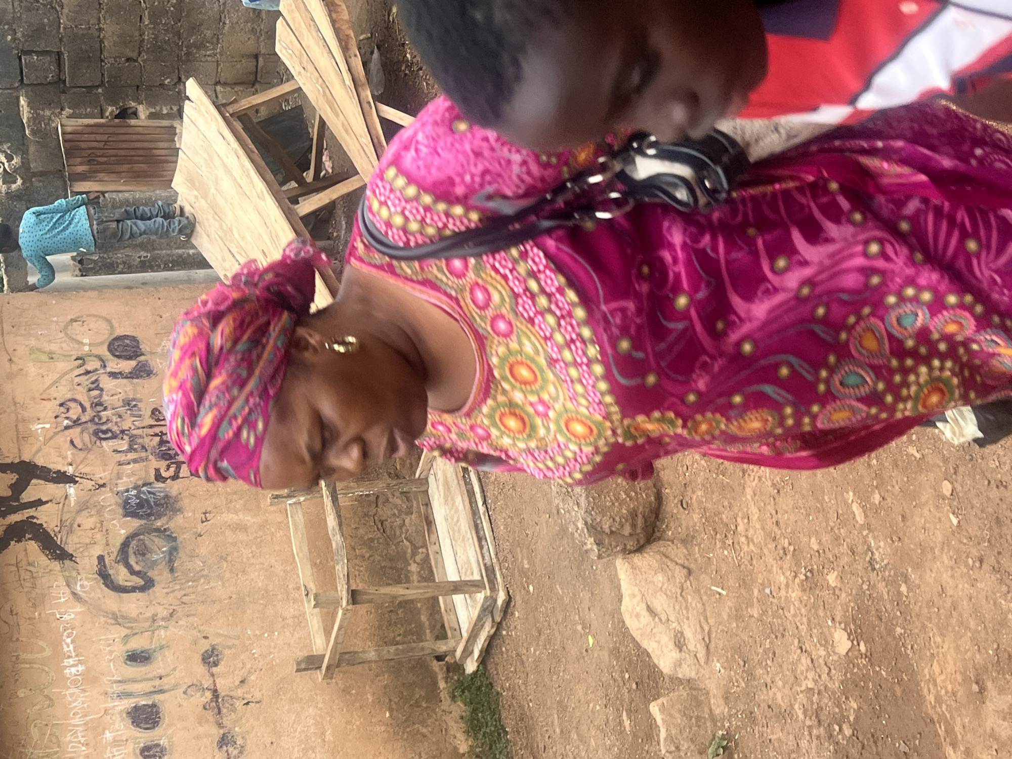 The image, rotated 90 degrees clockwise, depicts an outdoor scene in Jos, Nigeria, likely during the daytime. The setting features a dusty, reddish-brown earthen ground with scattered rocks and sparse vegetation. In the background, there's a wall made of rough concrete blocks, adorned with black and white graffiti-like markings, including a distinct 'S' and other unreadable script. Wooden planks and timber structures are visible throughout the scene, suggesting construction materials or makeshift arrangements.
In the immediate foreground, a woman with dark skin is prominent, dressed in a vibrant, patterned fuchsia dress with yellow, green, and orange designs. She wears a matching patterned headscarf and gold hoop earrings. A black shoulder bag with silver hardware rests on her shoulder. Her head is tilted downwards, and her expression is not clearly visible. Partially visible above her, to the right, is the head and shoulder of another person, possibly a child, wearing a red and white patterned garment, also looking downwards.
Further into the background, to the left, a third individual, likely a child or small adult, is seen bending over. They are wearing a light blue long-sleeved shirt with white polka dots and blue jeans. Near them, a small, simple wooden frame structure stands on the ground.
The scene appears to capture a candid moment in a bustling, potentially informal, urban or semi-urban environment. No specific activity is clearly discernible; the individuals seem to be going about their daily routines.