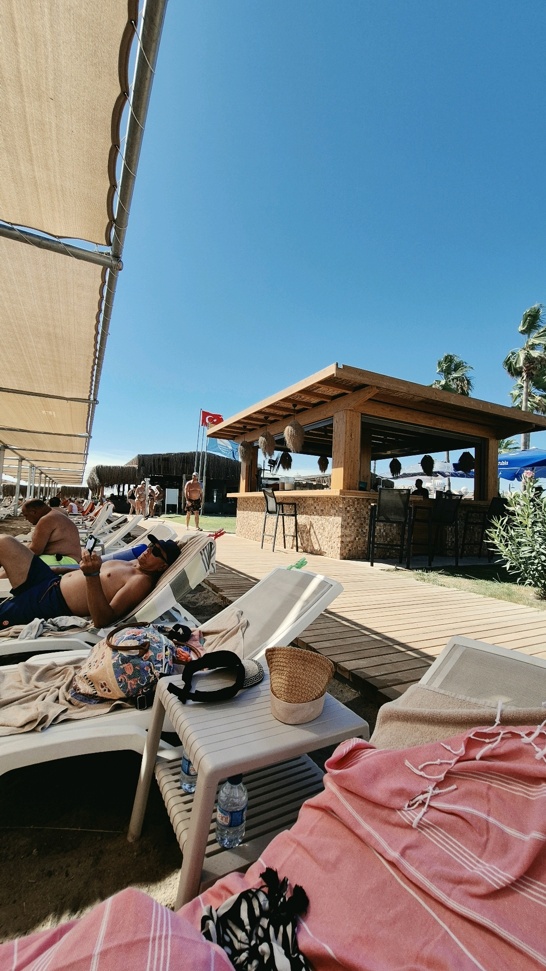 This is a full shot of a beach scene in Serik, Turkey, on a sunny day. In the foreground, several white lounge chairs with towels are arranged on a wooden boardwalk. On one of the chairs, a person is lying down wearing a black cap, sunglasses, and blue shorts, holding a smartphone. A small side table next to the chair holds a straw hat, a bag, and two bottles of water. A pink towel is draped over another lounge chair in the lower right corner. In the middle ground, there is a wooden beach bar with a thatched roof and seating area. Two Turkish flags are visible on poles behind the bar. Several people are scattered throughout the beach, some walking and some relaxing on lounge chairs. Palm trees and other vegetation are visible in the background. The sky is clear and blue.