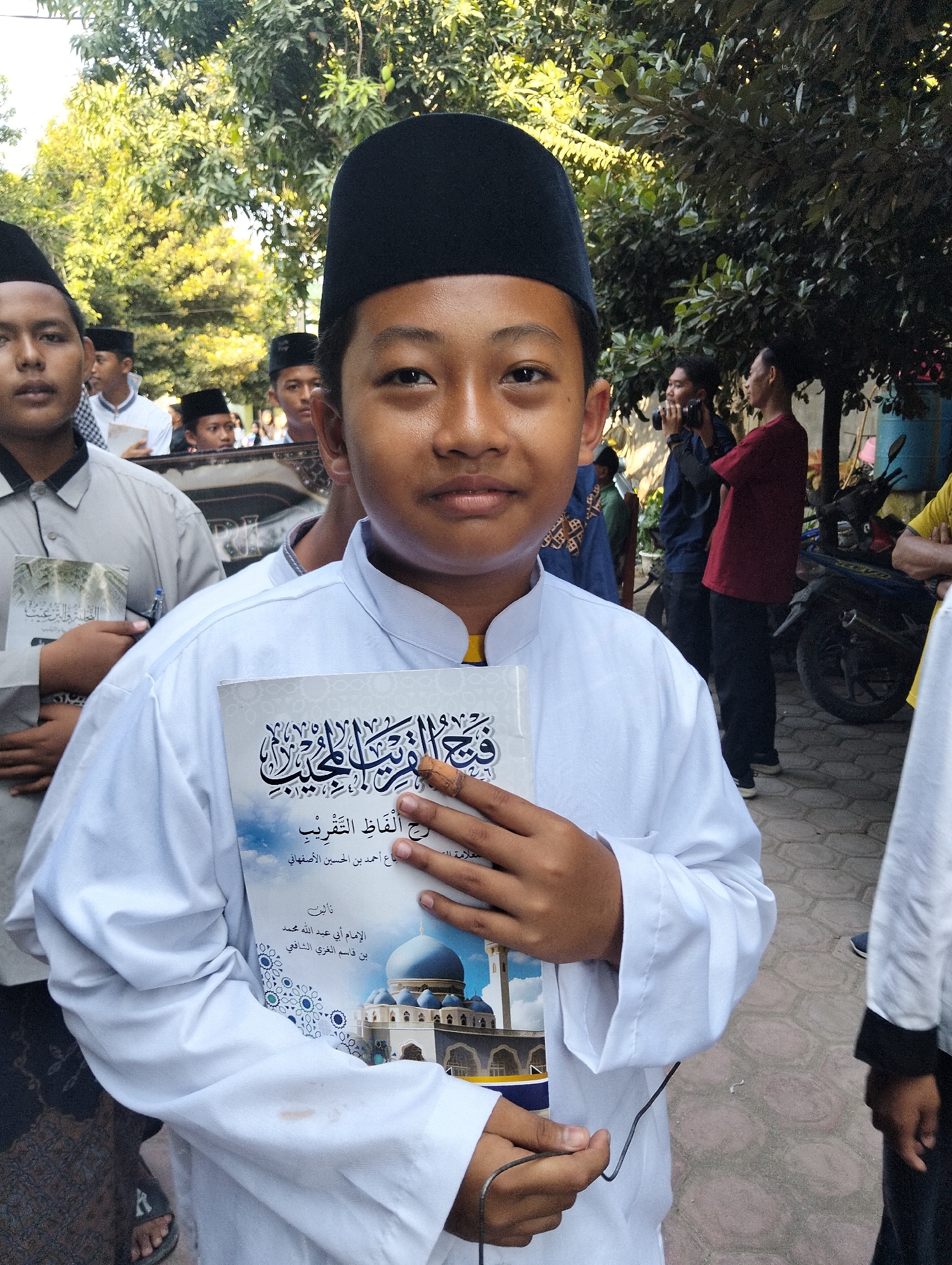 In Kranji, Indonesia, a young boy, likely a student, stands centrally, holding a book with Arabic script and a depiction of a mosque. He wears a white robe and a black, brimless hat. Behind him, other individuals, some also wearing similar attire, are visible amidst trees and paved ground. The atmosphere suggests an outdoor gathering, possibly related to a religious or educational event.