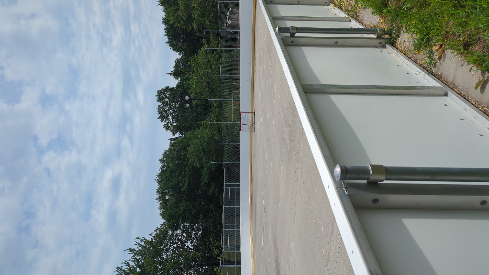The image shows an outdoor concrete hockey rink enclosed by a low white barrier and a chain-link fence. A red hockey net is visible on the rink in the distance. Trees form a backdrop behind the fence, and a cloudy sky is overhead. The scene appears to be located in Montreal, Canada, likely a public park or recreational facility, given the amenities present. There are no people or animals visible in the image, and no specific actions or interactions are depicted. The environment is an outdoor, man-made sports facility surrounded by nature.