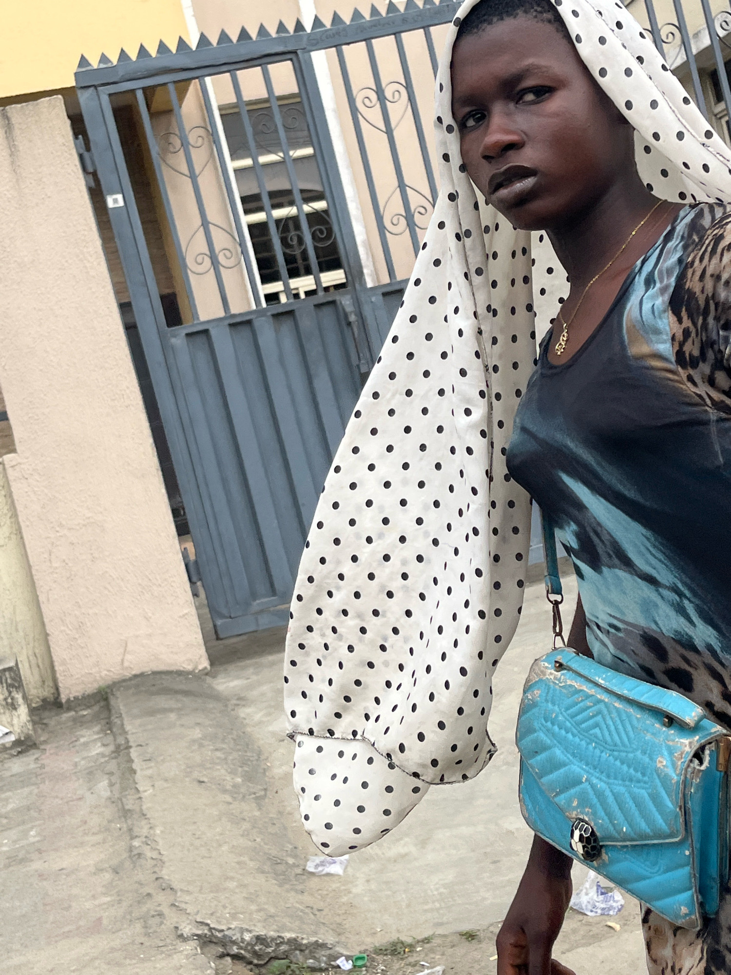 A young woman wearing a white headscarf with black polka dots and a patterned dress stands on a concrete walkway. She holds a distressed blue handbag with a snake-shaped clasp. Behind her is a gray metal gate with ornate scrollwork and pointed finials, which appears to be the entrance to a property. The building to the left of the gate is a light beige color. The ground is littered with small pieces of debris. The overall scene suggests a street or exterior setting within the city of Ado, Nigeria.