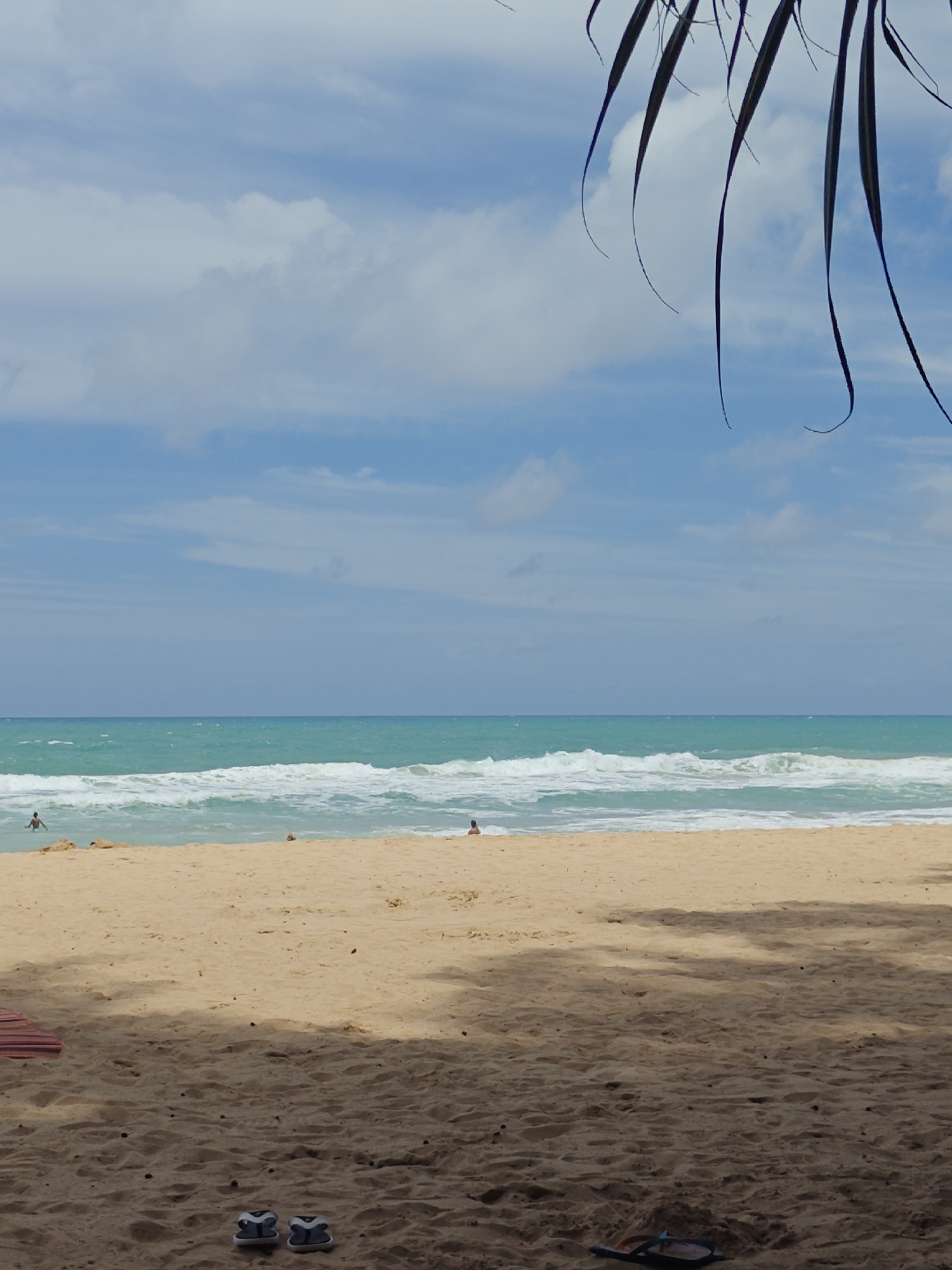 An outdoor, eye-level, medium shot captures a serene beach scene in Sakhu, Thailand. The image is dominated by the natural elements of sand, sea, and sky. The foreground features a sandy beach with subtle shadows cast from unseen palm trees. Two pairs of flip-flops rest on the sand in the lower portion of the frame. A striped beach mat is partially visible on the left side of the foreground. In the middle ground, the golden sand gives way to the turquoise ocean, with gentle waves rolling towards the shore. Several individuals are scattered across the water and beach. One person is visible in the shallow water on the left, while another appears to be sitting further down the beach on the right. The horizon is a sharp line where the sea meets the sky, which is a clear blue dotted with soft, white clouds. A few palm fronds hang from the top edge of the frame, framing the scene.