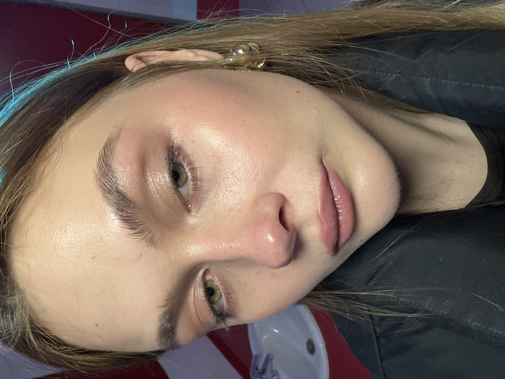 A close-up portrait of a fair-skinned young woman with blonde hair. She has light green eyes, defined eyebrows, and glossy pink lips. She is wearing a gold earring. A sink is visible in the lower center of the frame. A black jacket is seen on her right side. The background appears to be a room with red walls. There is no information to suggest the location is in Krasnoyarsk, Russia.