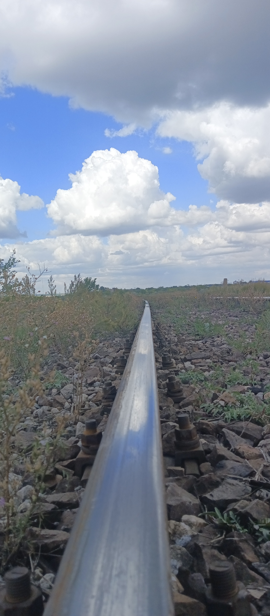 A low-angle, vertical shot captures a single railroad track stretching into the distance under a partly cloudy sky. The track, made of polished steel, is the dominant subject in the foreground, reflecting the sky. It is laid on a bed of rough stones and gravel, with some scattered green vegetation and dry weeds growing around it. The tracks curve slightly to the right as they recede, disappearing into the hazy horizon. In the far distance, a faint industrial structure is visible on the right side. The sky is a mix of bright blue patches and large, fluffy white and gray clouds, suggesting a dynamic weather condition. The image appears to have been taken outdoors during the daytime. The location is identified as being in Karaganda, Kazakhstan.