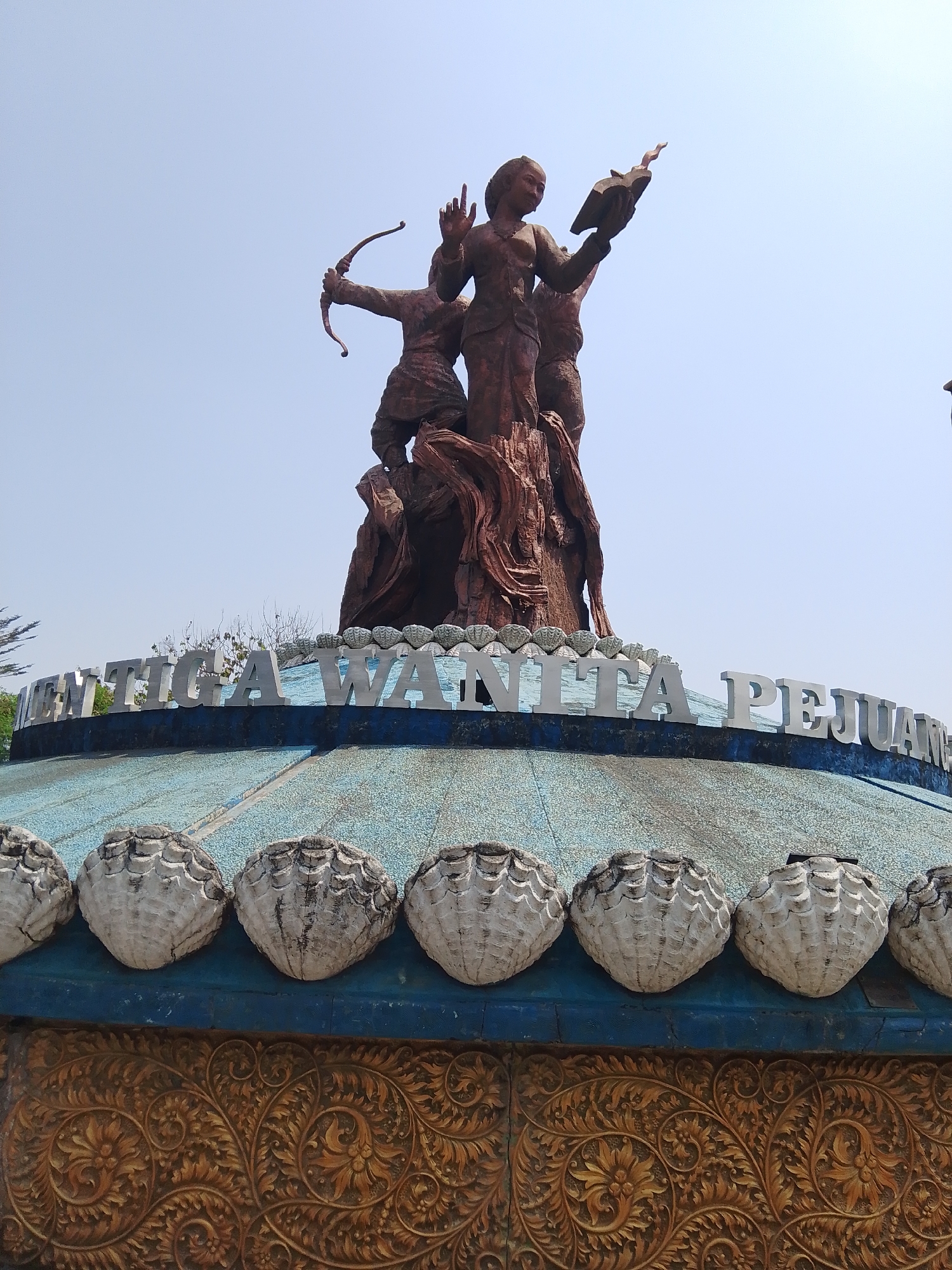 The image shows a bronze statue of three women, identified by the text on the structure below as "TIGA WANITA PEJUANG," which translates to "Three Warrior Women." One woman is depicted drawing a bow and arrow, another is holding an open book with flames emanating from it, and a third stands between them. The statue is positioned atop a circular structure with a blue roof adorned with a row of white, shell-like ornaments. Below this, a golden, ornate relief features intricate floral and scrollwork patterns. The statue is seen against a clear, pale blue sky. The location is specified as Tahunan, Indonesia.