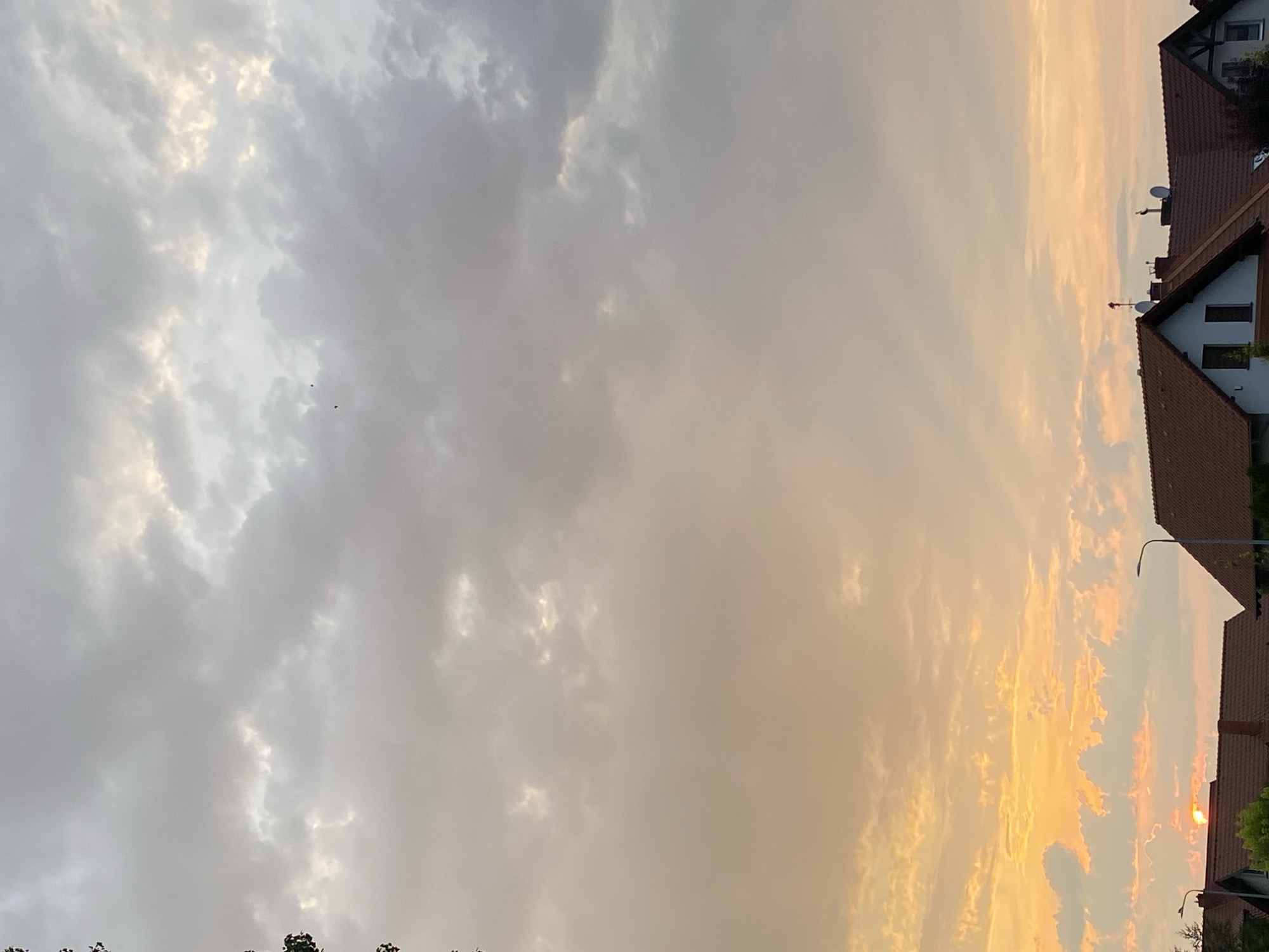 The image depicts a sunset over a residential area in Buk, Poland. The sky is filled with dramatic clouds, with the lower portion illuminated by the warm hues of the setting sun, transitioning to cooler grays and whites towards the top. A sliver of the sun is visible on the horizon, peeking through the clouds above the rooftops. Several houses with terracotta roofs are visible in the foreground, with streetlights and antennas on some of the roofs. The scene appears to be a peaceful evening in a suburban setting.