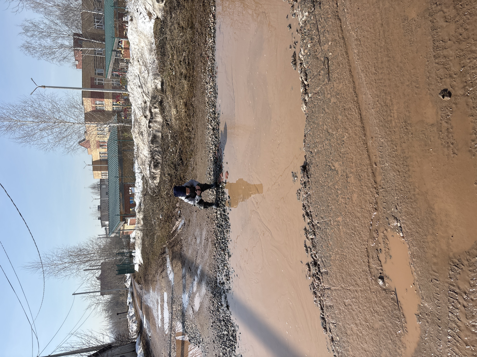 A person stands at the edge of a water-filled pothole in a street. The pothole is extensive, occupying a large portion of the roadway. The road surface adjacent to the water is broken and uneven. The side of the street opposite the person has melting snow and buildings can be seen in the background, including houses with green roofs and brick walls. Bare trees are interspersed among the buildings. The sky is a pale blue. Wires run across the sky in the upper left of the image. This is in Ferma, Russia.