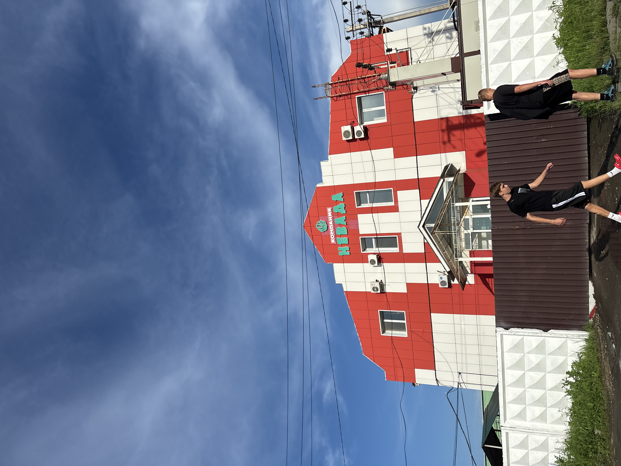 Two young men walk past a red and white striped building in Blagoveshchensk, Russia, under a partly cloudy blue sky. The building has "КОМПАНИЯ НИКА" written in green letters across its upper facade, along with a green logo. There are several windows and air conditioning units visible on the building. A white concrete fence with a geometric pattern borders the foreground, with a brown metal fence to its right. Overhead power lines crisscross the sky. The ground in front of the fences appears wet and muddy in places. One man on the left wears a black t-shirt, black shorts with white stripes, and red sneakers. The man on the right wears a black t-shirt, plaid shorts, and dark athletic shoes.