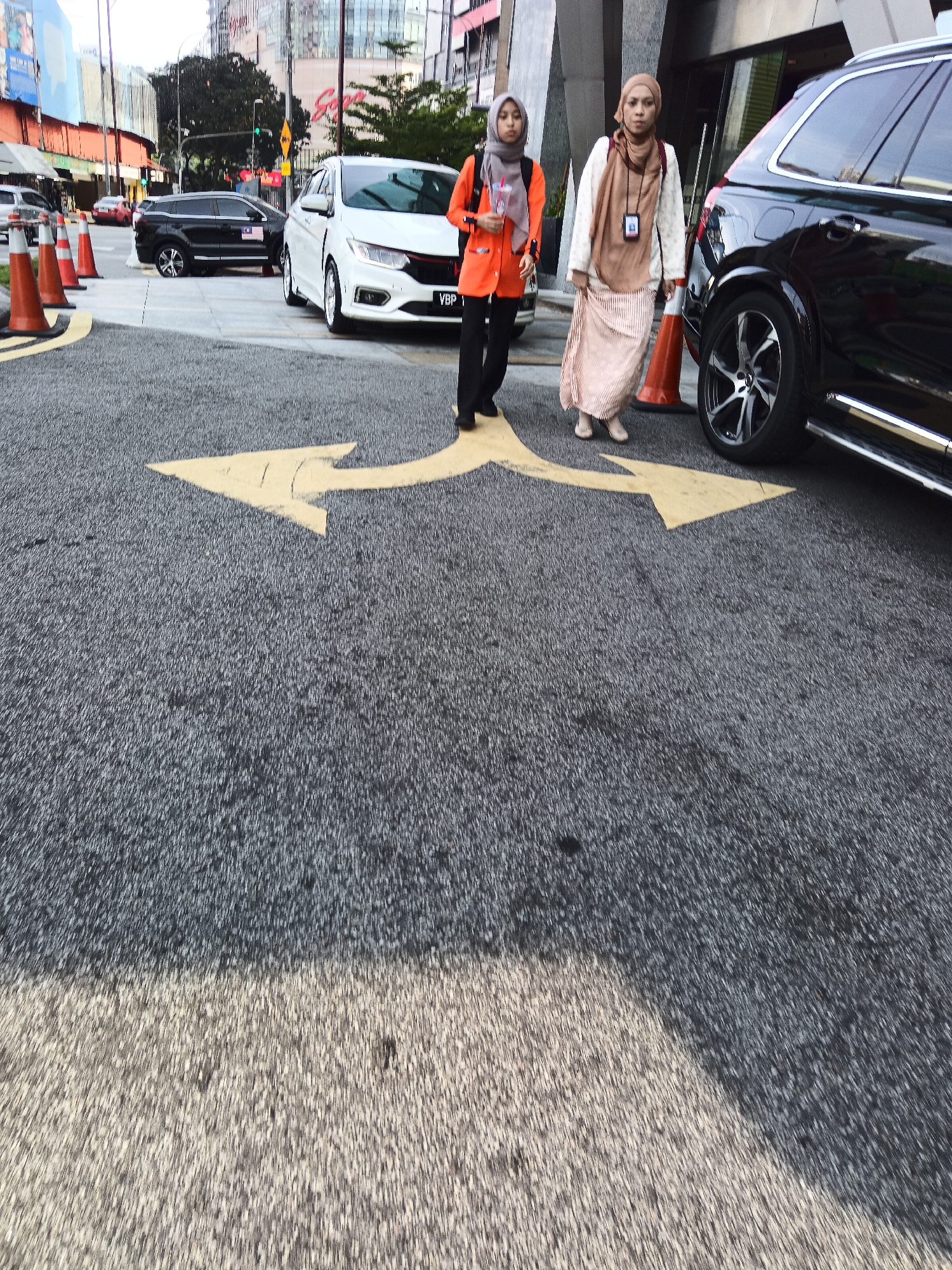 Two women are walking on a street in Kuala Lumpur, Malaysia. The woman on the left is wearing an orange jacket and a grey hijab. She is carrying a backpack. The woman on the right is wearing a white sweater, a brown hijab, and a long skirt. She is holding a lanyard with an ID card. They are walking on an asphalt road with yellow arrows painted on it, indicating a turn. In the background, there are cars, including a white Honda and a black SUV. There are also buildings, traffic cones, and a billboard. The scene appears to be a street or a parking lot in an urban area.