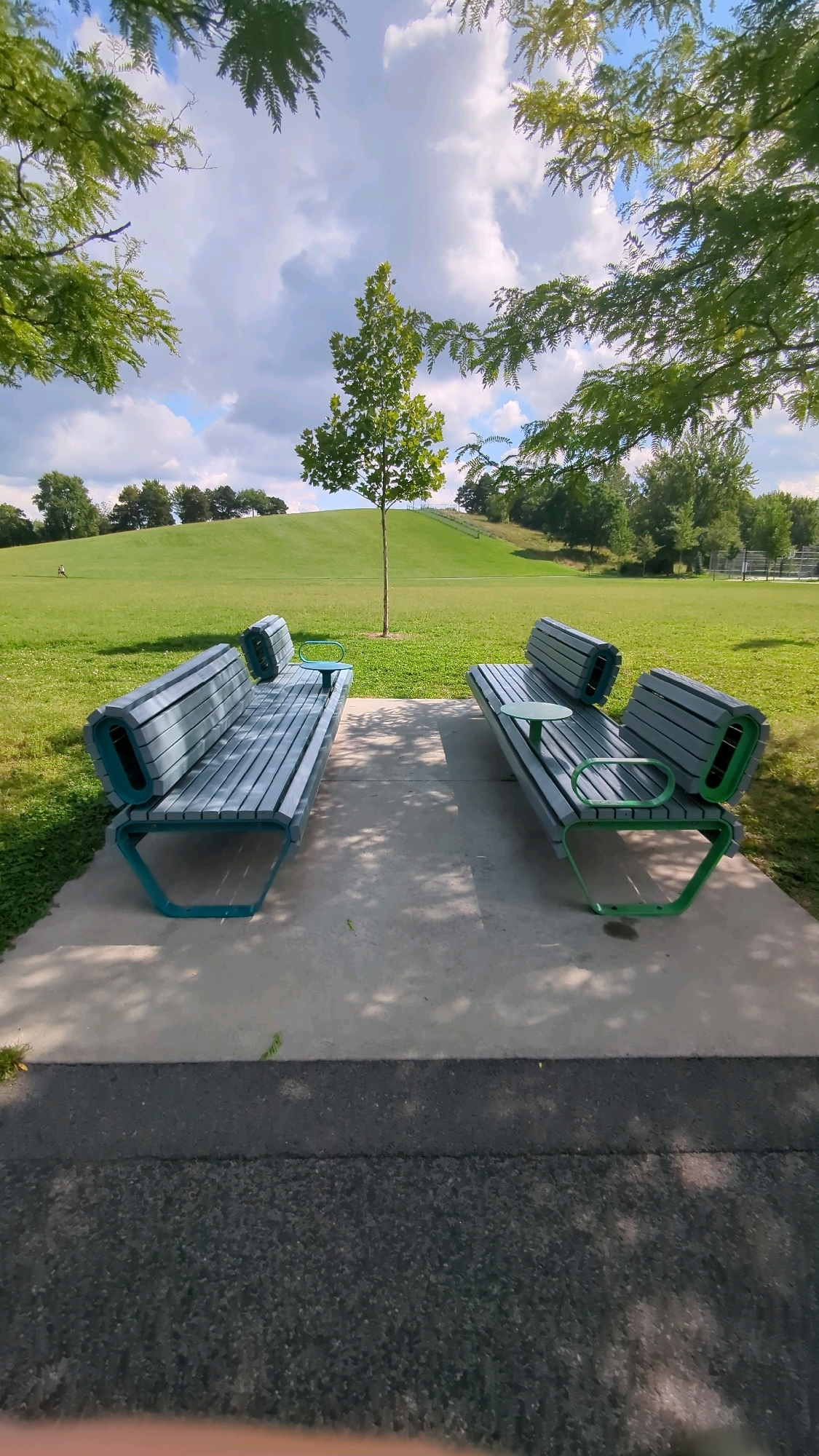 The image depicts two contemporary park benches with teal frames and gray slatted seating, placed on a concrete patio in an urban park. The benches are positioned facing each other, separated by a small distance. To the left of the benches, a strip of green grass borders the patio. Beyond the patio, a large, gently sloping green hill dominates the background. A solitary, slender tree stands on the hill, with a single person walking in the distance. The park is surrounded by various trees with lush green foliage, framing the scene under a partly cloudy sky. The overall impression is of a serene and well-maintained public space. The context of Montreal, Canada, suggests this scene is likely from a park within the city.