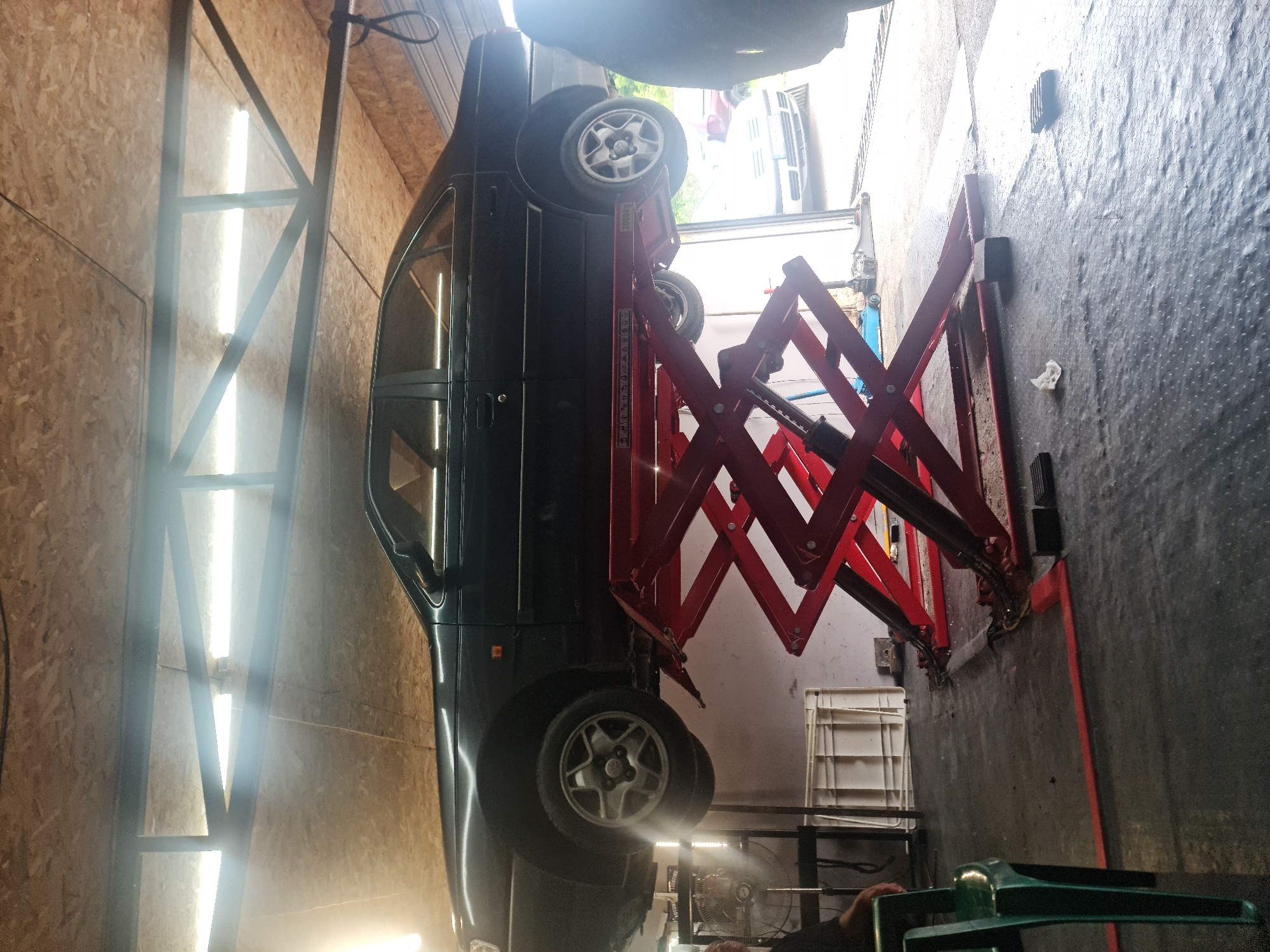An indoor garage or workshop in Buzovgrad, Bulgaria, features a dark-colored car elevated on a bright red scissor lift. The car, possibly an SUV or minivan, is seen from its side, suspended in mid-air with its alloy wheels clearly visible. The red hydraulic lift's crisscrossing arms support the vehicle.
The left wall of the garage is constructed from light-colored wooden panels, likely OSB or plywood, featuring long, bright fluorescent lights and dark metal support beams. The floor beneath the lift and car is a dark, textured rubber mat covering concrete.
Various other objects are scattered around the workspace, including a white plastic crate or shelf unit, a black stand, and a small white crumpled rag on the floor. A person's hand is visible in the lower right corner, near a green object, possibly a tool or piece of equipment. Further back, a fan can be discerned.
The bright artificial lighting from the fluorescent tubes suggests an active workspace, regardless of the time of day. Some text, possibly "BUDAPES...", is visible on the side of the red scissor lift, likely indicating a brand or origin. The scene depicts a vehicle undergoing maintenance or inspection.