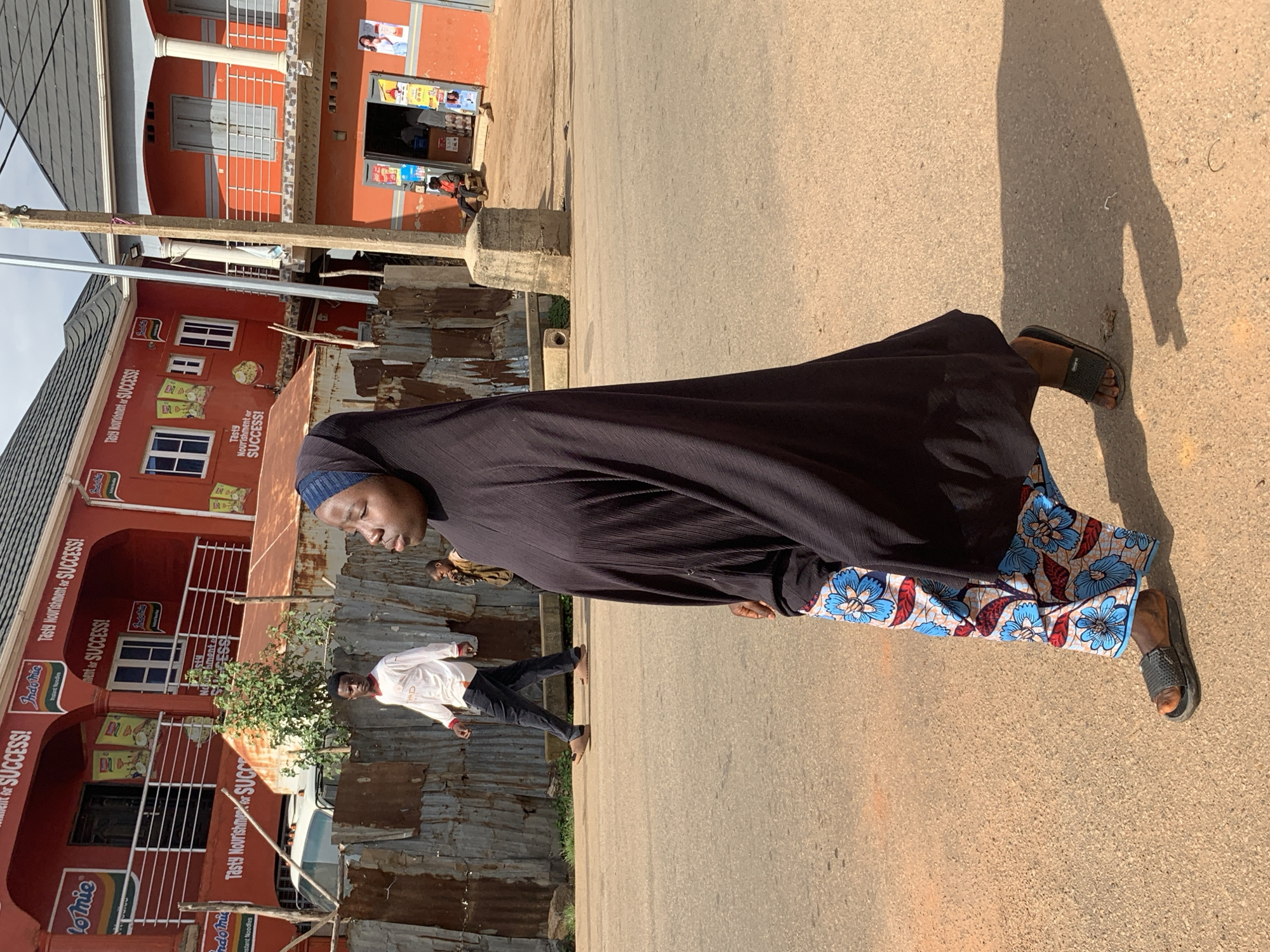 In the city of Jos, Nigeria, a woman wearing a black hijab and a patterned skirt walks across a paved road. Her skirt features a vibrant blue and red floral print with a white background. She is wearing dark-colored slip-on sandals. In the background, a young man in a white long-sleeved shirt and dark pants walks away from the camera. Further in the background, a young boy is visible. To the left, a corrugated metal fence separates the road from buildings. The buildings are painted orange and red, with some displaying advertisements for Indomie noodles and the slogan "Tasty Nourishment for SUCCESS!". A concrete pillar stands in the middle of the road. The sky is overcast.