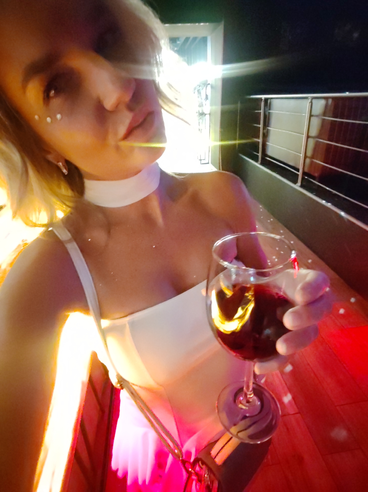 A woman with blonde hair, wearing a white dress and a white choker necklace, is holding a glass of red wine. She has white dots applied to her cheekbones. She is holding a yellow straw in her mouth. The scene appears to be outdoors at night, possibly on a balcony or patio, with a railing visible in the background. Red and bright yellow lighting illuminates the scene. There is no visible text. The overall mood seems festive or celebratory.