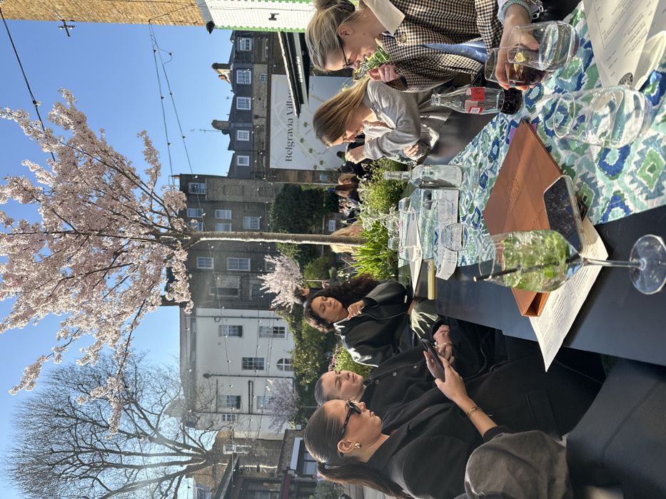 The scene depicts an outdoor dining area on a clear, sunny day in Belgravia, London, United Kingdom, identifiable by a "Belgravia Village LONDON" sign in the background. The environment includes historic brick and white buildings, a prominent cherry blossom tree in full bloom, a bare deciduous tree, and overhead string lights.
In the foreground, six women are seated at a long, dark rectangular table covered partially by a blue and white patterned table runner. From left to right:
1. A woman with dark hair tied back, wearing sunglasses and a dark jacket, is observed holding and looking at a black smartphone. She wears gold earrings.
2. A woman with dark hair, wearing a dark top, is seated, facing forward.
3. A woman with dark, curly hair, wearing a dark leather-like jacket, is looking down.
4. A woman with blonde hair, wearing a light grey long-sleeved top, is looking down and to her left.
5. A woman with blonde hair, wearing glasses and a brown and white checkered jacket with a light collar, is looking to her right.
6. The rightmost woman is partially visible, holding a glass containing a dark liquid with a lime wedge.
On the table, various items are present: a tall glass containing a green cocktail with mint and a straw, a black smartphone, a brown menu or folder (partially legible as "Home House"), multiple empty and filled drinking glasses, a clear water bottle, a red Coca-Cola bottle, and paper menus/placemats. The women appear to be engaged in social interaction.