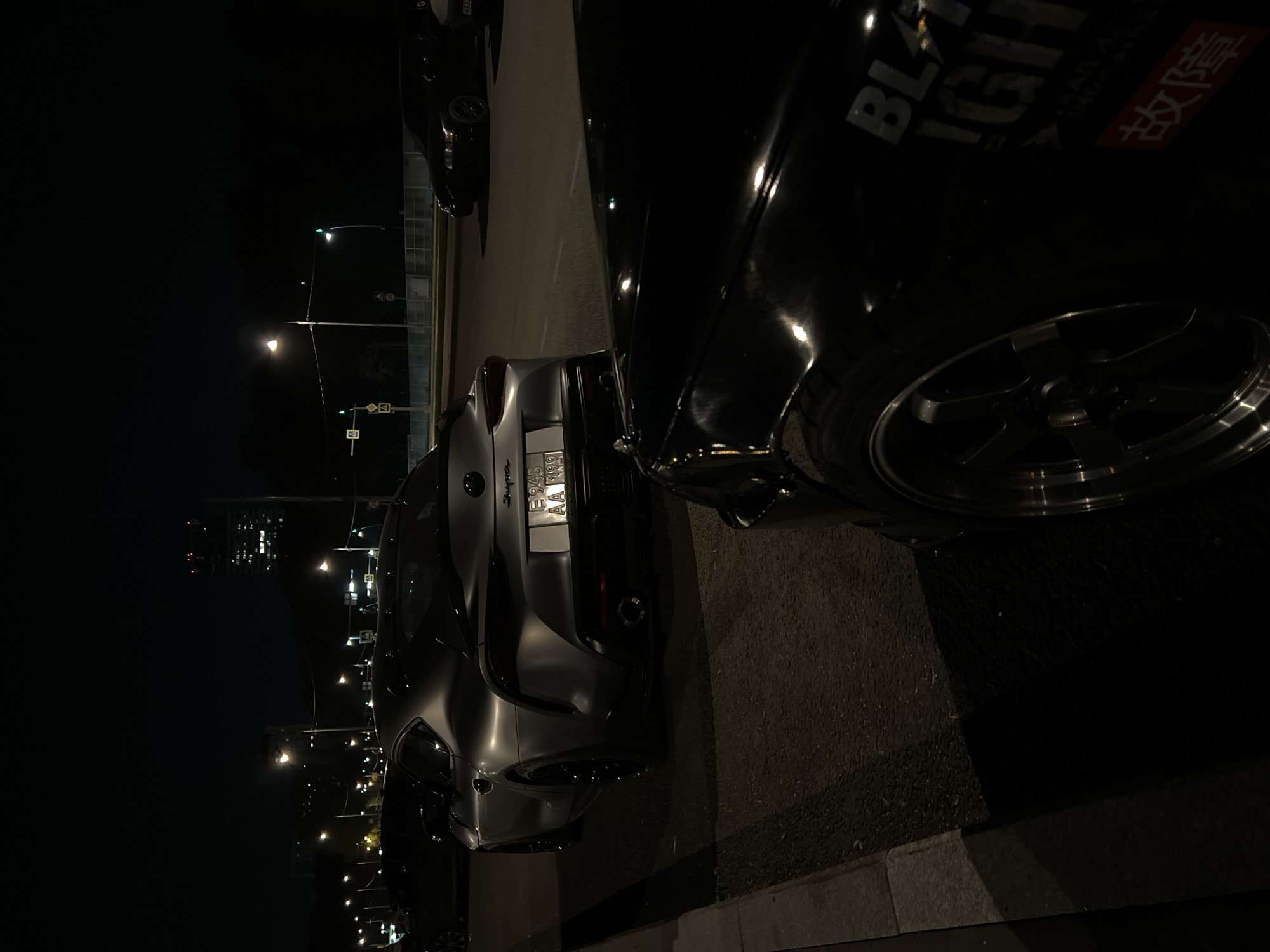 The image shows a dark parking lot at night in Khimki, Russia. Streetlights are visible in the background, along with other buildings. Three cars are parked in a row. The closest car to the camera is black with white lettering that includes the word "BLAZING". The second car is a silver Toyota Supra with the license plate "E945AA 190". The third car is partially visible and black.