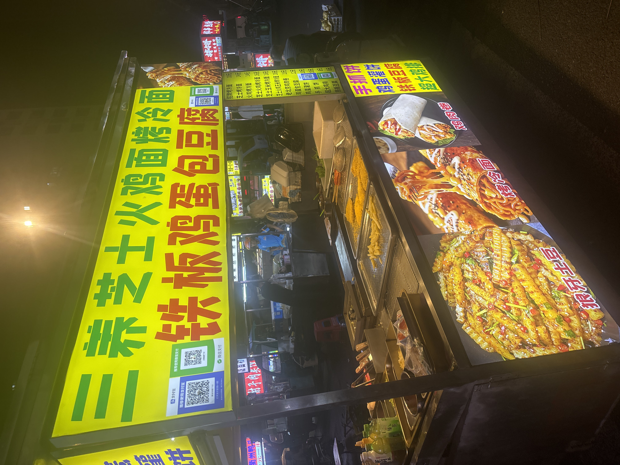 An outdoor food stall in Suzhou Industrial Park, China, at night, is illuminated by a bright yellow neon sign displaying menu items. The sign features large Chinese characters and smaller text listing various food options like "three cheese cheese ramen," "iron plate chicken egg tofu," and "spicy cold noodles." Two QR codes for mobile payment are prominently displayed. The stall itself has stainless steel trays filled with food, including french fries and what appears to be fried chicken. A close-up image of loaded french fries is displayed on the front of the stall. In the background, a person in a blue shirt stands behind the counter, and other individuals can be seen in the dimly lit street.
