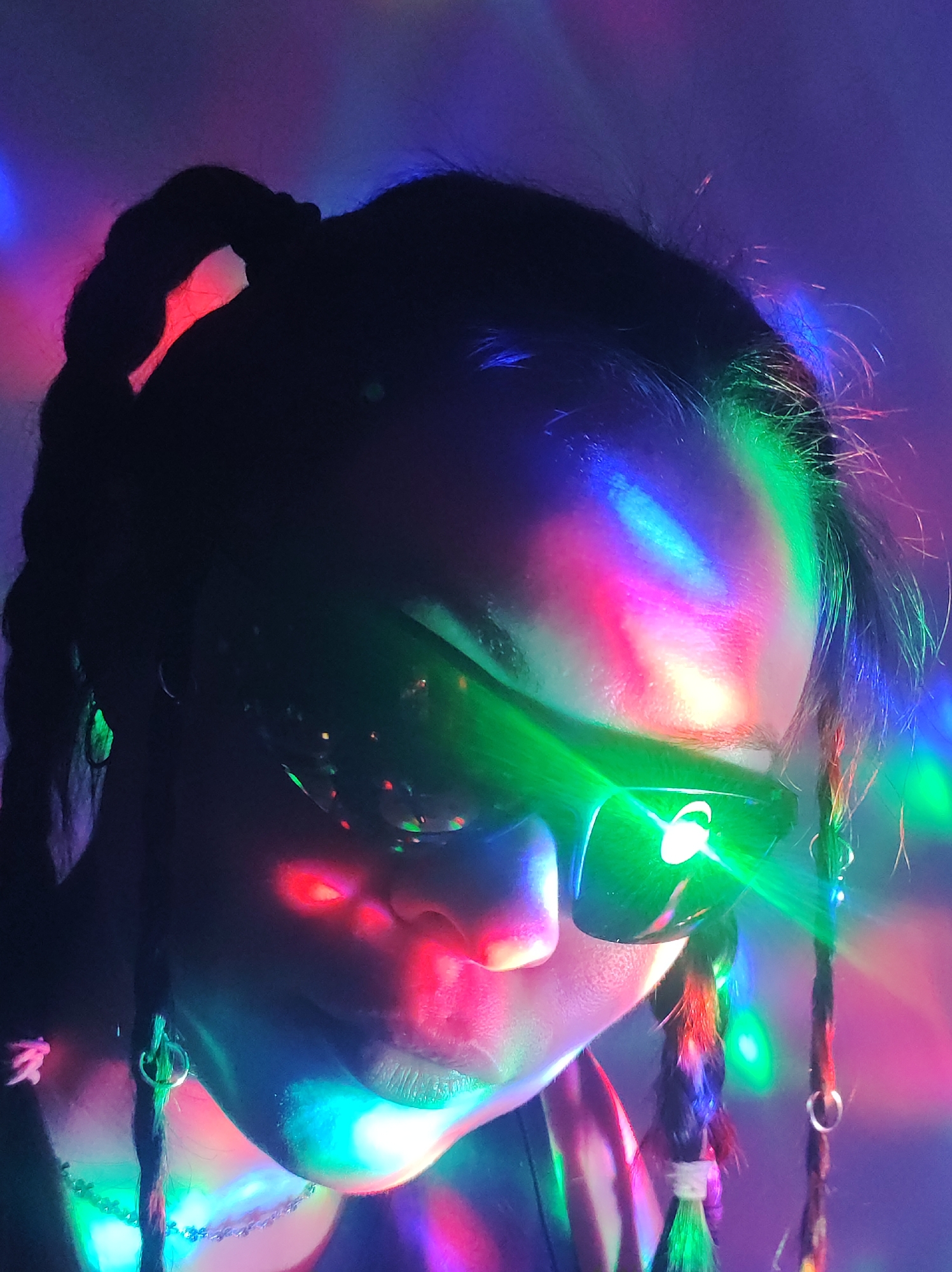 A close-up, portrait-style photograph features a person with dark hair, possibly styled in braids or dreadlocks, wearing dark, reflective sunglasses. The scene is illuminated by a vibrant, multi-colored light display, creating a dynamic and energetic atmosphere with hues of red, green, blue, pink, purple, and yellow.
The person's face is partially visible below the sunglasses, with their forehead and temples illuminated by streaks and patterns of various colors. The reflective sunglasses intensely catch and refract the colorful lights, creating bright highlights and green light beams across the lenses. The lower part of the face, including the nose, lips, and chin, is also bathed in the shifting colored light.
Hair braids or dreadlocks are visible, some adorned with small rings or colorful accents. An earring can be seen on the visible left ear, and a thin, possibly beaded, necklace is faintly discernible around the neck area. The background is indistinct, suggesting an indoor setting with low ambient light, enhanced by special effect lighting, reminiscent of a party or club environment. The overall impression is one of cool mystery and vibrant energy. No text is visible in the image.