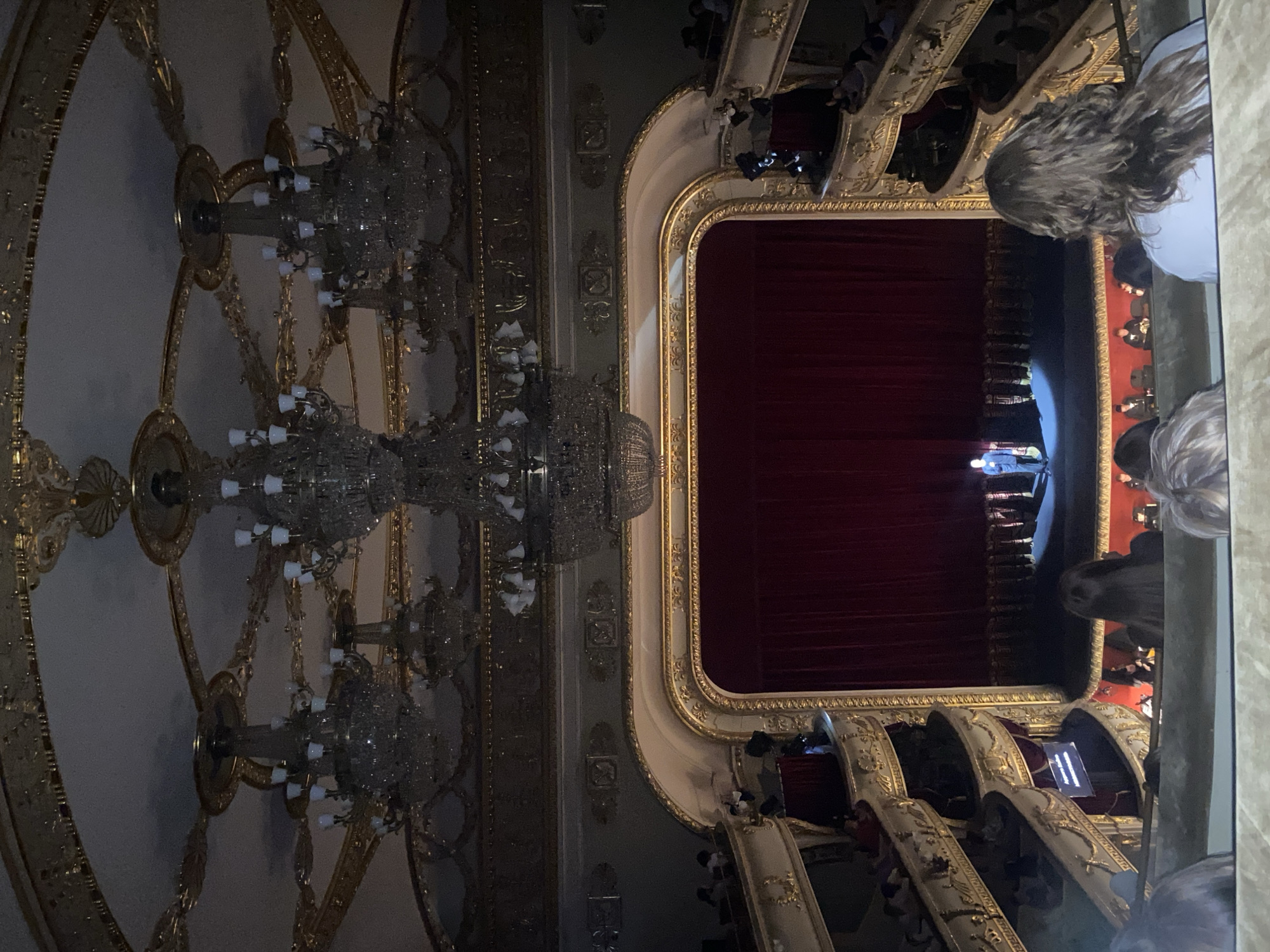 The image shows the interior of the Yekaterinburg State Academic Opera and Ballet Theater in Yekaterinburg, Russia. Three large crystal chandeliers illuminate the ornate ceiling and the walls decorated with gold trim. A large stage framed with gold and cream-colored molding is visible. A red curtain hangs on the stage. People are seated in the theater boxes, looking towards the stage. There appear to be performers on the stage.