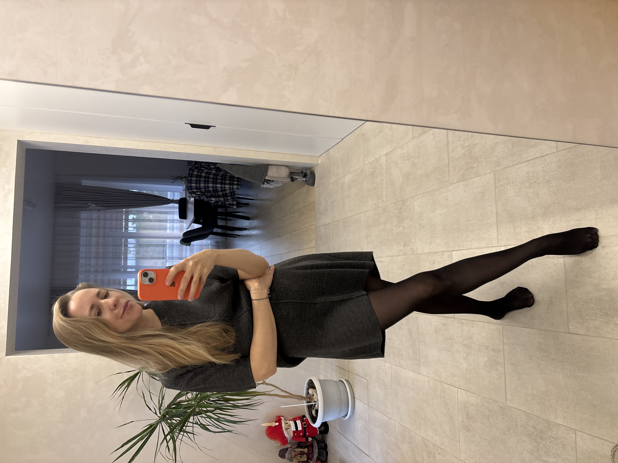 A woman is taking a selfie in an indoor environment. She is blonde with long hair and wearing a gray dress and black tights. She is holding an orange-cased mobile phone.
Behind her is a doorway leading to another room with visible furniture and curtains. A potted plant and decorative figures are near her. The floor is tiled. Based on previous knowledge, the location is Chaikovsky, Russia.