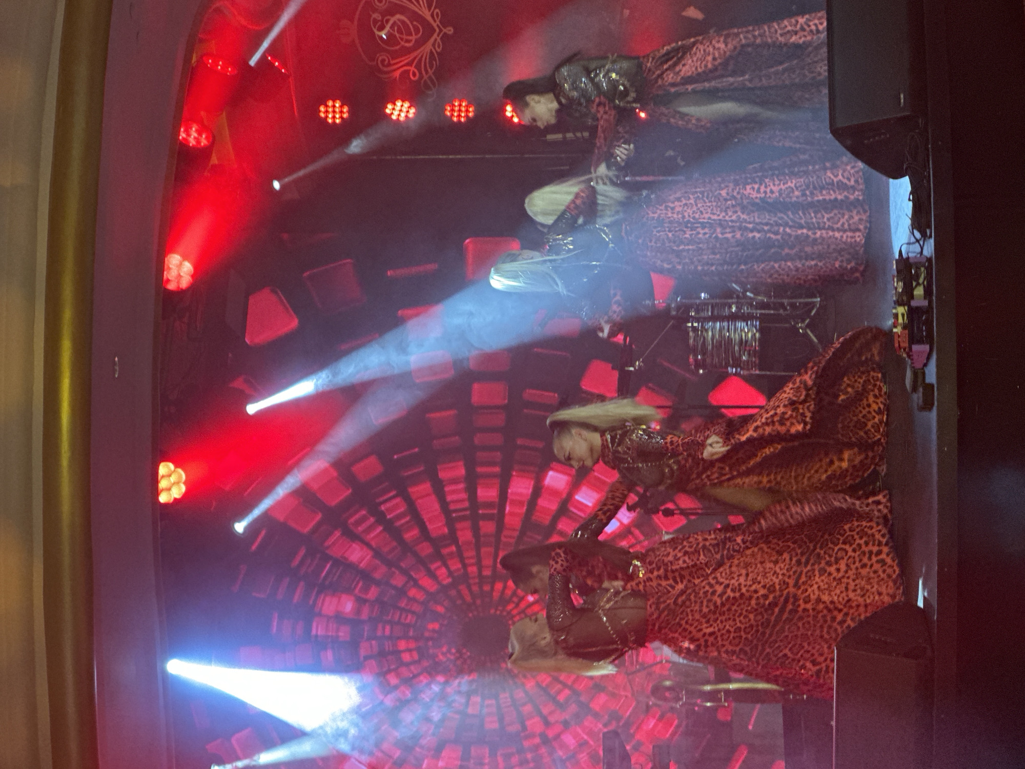 A group of female performers are on a stage. They are wearing matching leopard print dresses with high slits and sparkly tops. They have their hair pulled back in ponytails. Some of the performers are holding each other. The stage is lit with red and white lights, and there is a large screen behind them with a red, geometric pattern. There are speakers on the stage and musical equipment. The performance is taking place in the city of Estosadok, Russia.