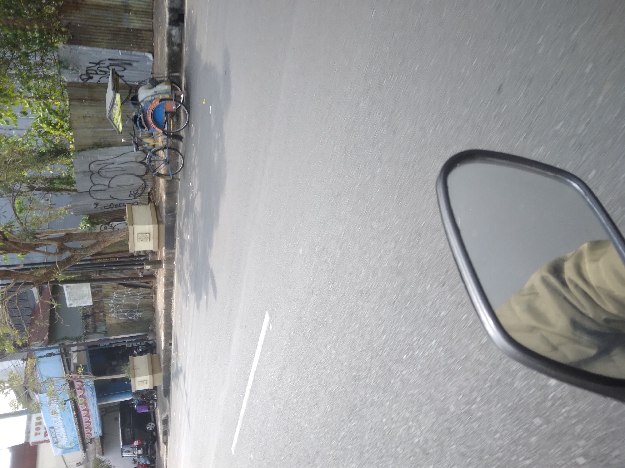 The image depicts a street scene in Purwokerto, Indonesia. On the left side of the frame, a black car is parked in front of a building with a blue facade and signage that reads "TOKO" and "KARYA SANTRI". A tricycle, known locally as a "becak", is stationary on the right side of the road. The becak has a blue seat and a yellow canopy. The road surface is asphalt, and a white line marks the lane. In the foreground, a motorcycle's rearview mirror is visible, reflecting a person's arm and shoulder wearing a light brown jacket. The background includes a corrugated metal fence with graffiti and several trees with green foliage. The overall environment appears to be an urban street with commercial establishments and traffic.
