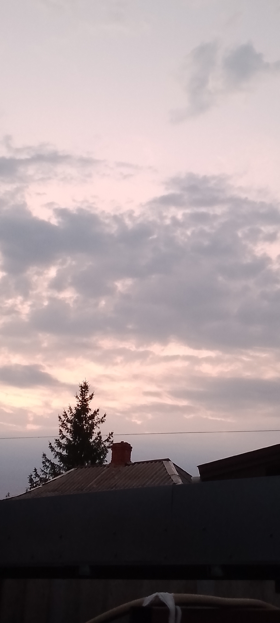 The image depicts a twilight sky with pale pink and grey clouds. In the foreground, silhouetted against the sky, is the roof of a building with a corrugated metal surface. A brick chimney with a red cap stands on the roof. To the left of the chimney, the dark silhouette of a tall evergreen tree is visible. In the far foreground, a dark, horizontal structure, possibly a fence or another building, spans the width of the image. The scene is consistent with a residential area at dusk in Belgorod, Russia, with no people or discernible activity present.