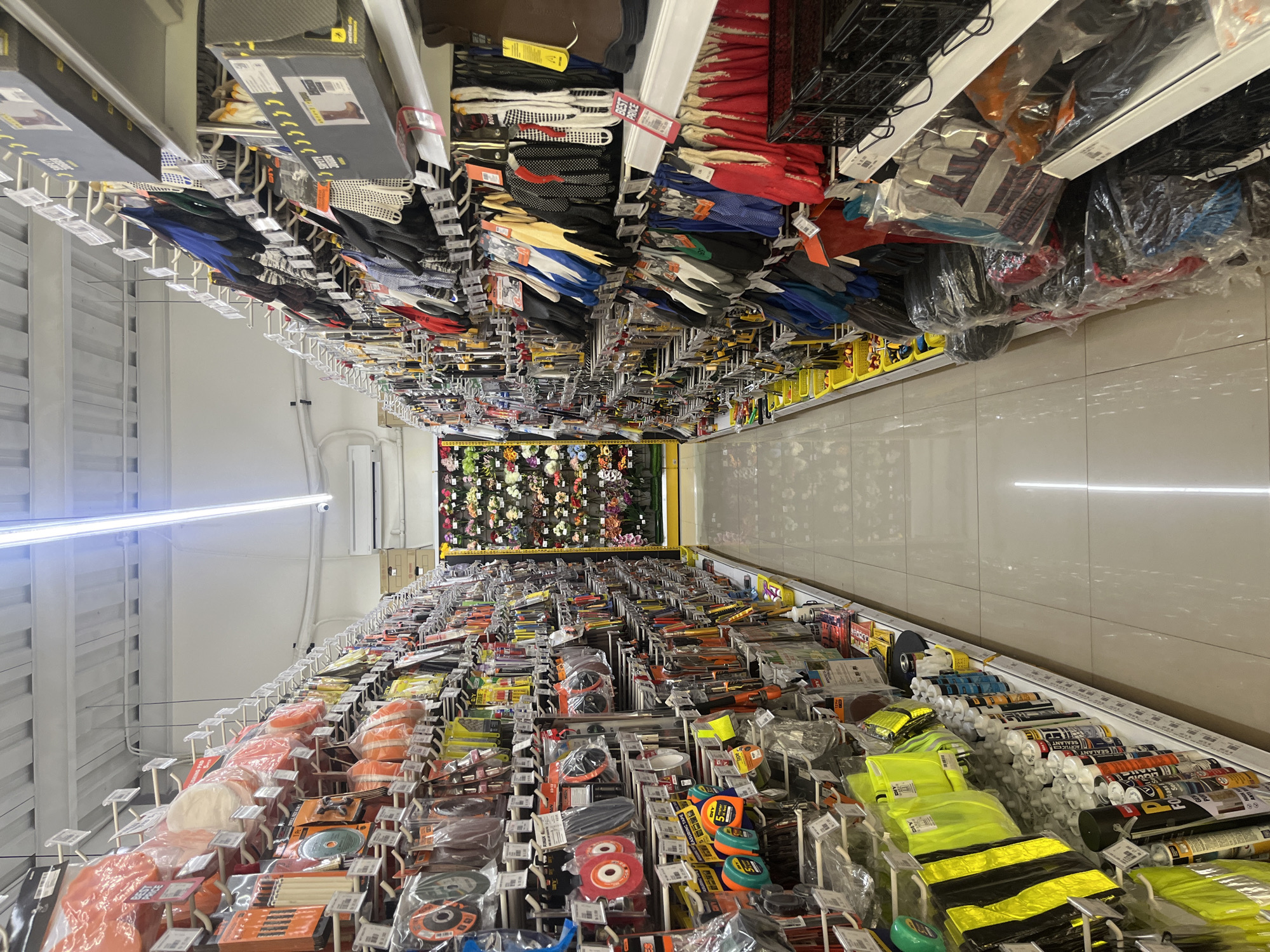 The image captures a long, brightly lit aisle within a hardware or home improvement store, viewed from one end looking towards the other. The perspective emphasizes the depth of the aisle, which is lined with tall shelving units on both sides, packed with various products. The floor is covered with large, light-colored tiles, and the walls are painted a uniform light tone, suggesting an organized commercial space. A long fluorescent light fixture runs along the ceiling, providing ample illumination.
On the left side of the aisle, a wide array of hardware items is displayed on hooks and shelves. These include numerous packaged tools, such as cutting discs, measuring tapes, and various small hand tools. Also visible are stacks of bright yellow and black reflective vests, and a section with tubes of sealants and adhesives, stacked horizontally. Many of these products are encased in clear plastic packaging.
The right side of the aisle features a large selection of gloves of different types, including work gloves, safety gloves, and possibly gardening gloves, hanging neatly from hooks. Above these gloves, some larger boxed items are arranged on shelves. The items here vary in color and material, showcasing a range of options.
Towards the far end of the aisle, a vibrant display of what appears to be artificial flowers or decorative plants adds a splash of color against the back wall, breaking the functional aesthetic of the hardware items.
No people are present in the image. The store appears clean, well-stocked, and orderly, indicating a typical retail environment during business hours. Visible text includes "BEST PRICE" on a red tag near some gloves, and various labels on the sealant tubes such as "SEALANT", "FLEXIBLE SEALANT", "LIQUID NAILS", and "FOAM SEALANT." Other partially visible text includes product names or brands like "SET", "SGS", and "MEASURING TAPE."
