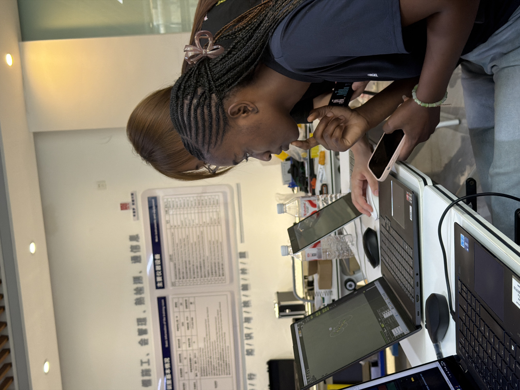 The image shows two women in a room with computers. The woman in the foreground, with dark skin and braided hair, is wearing a dark t-shirt and grey pants. She is looking at a phone in her hand and her other hand is resting on a laptop. She is wearing a green beaded bracelet. The woman in the background, with lighter skin and brown hair, is wearing a black t-shirt and glasses. She is looking at a laptop and her hand is on a mouse. There are two laptops, two computer mice, and two water bottles on the table. A whiteboard with Chinese text is visible on the wall in the background. The overall lighting suggests an indoor environment, possibly a classroom or office space. Based on the context of similar images, the location is likely within the city of Jiangjin, China.