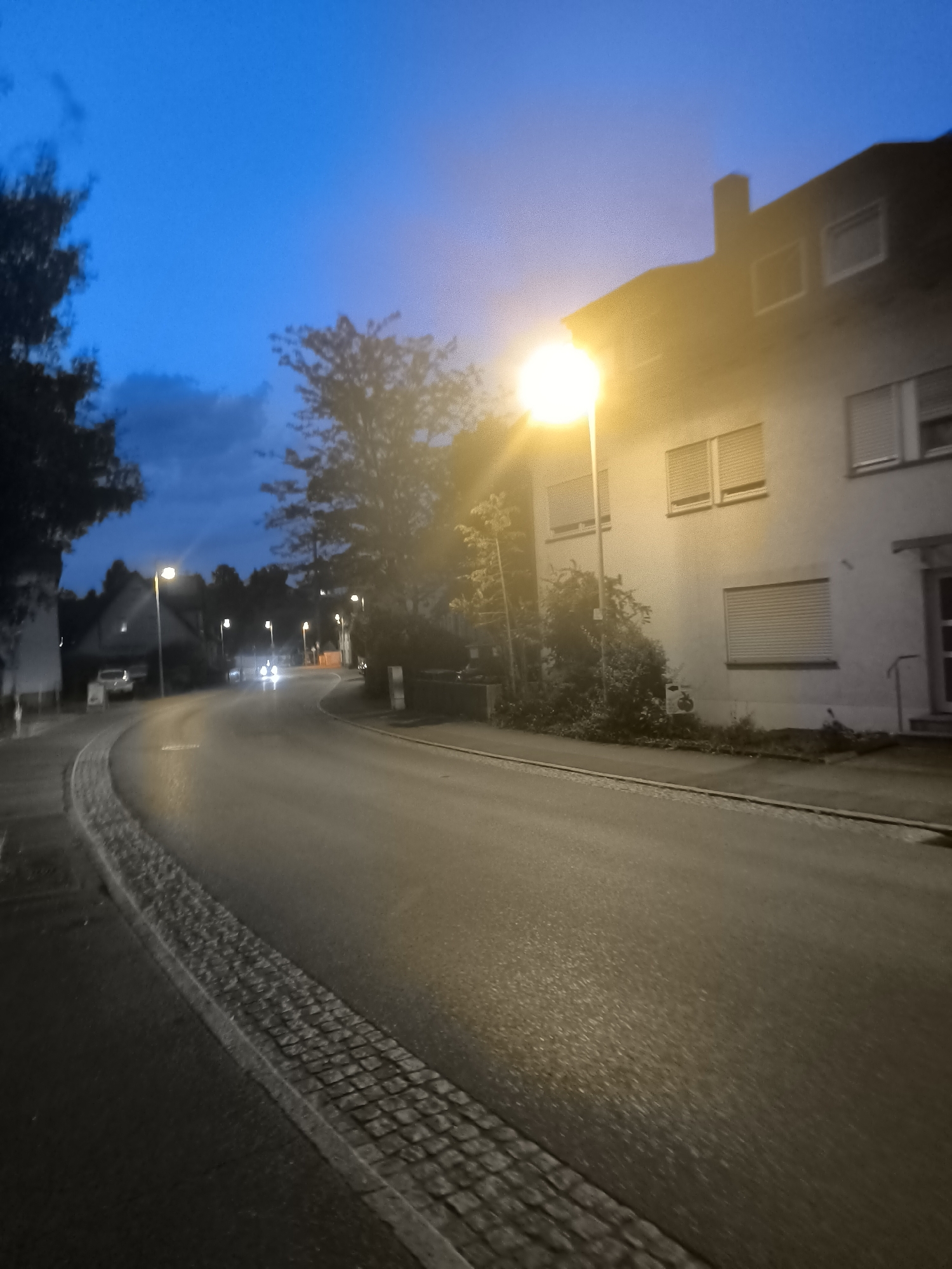 The image depicts a street scene at dusk in an unknown urban or suburban location. A paved road curves to the left, bordered by a cobblestone curb on the left side of the foreground. A street light illuminates the scene with a bright yellow glow from the right side of the image, casting light on a multi-story building with windows and shutters. Trees and bushes are visible along the side of the road and in front of the building. In the distance, the headlights of a vehicle are visible on the road, and several other street lights line the street. The sky is a deep blue with some clouds, indicating twilight. The overall impression is one of a quiet street at the end of the day.