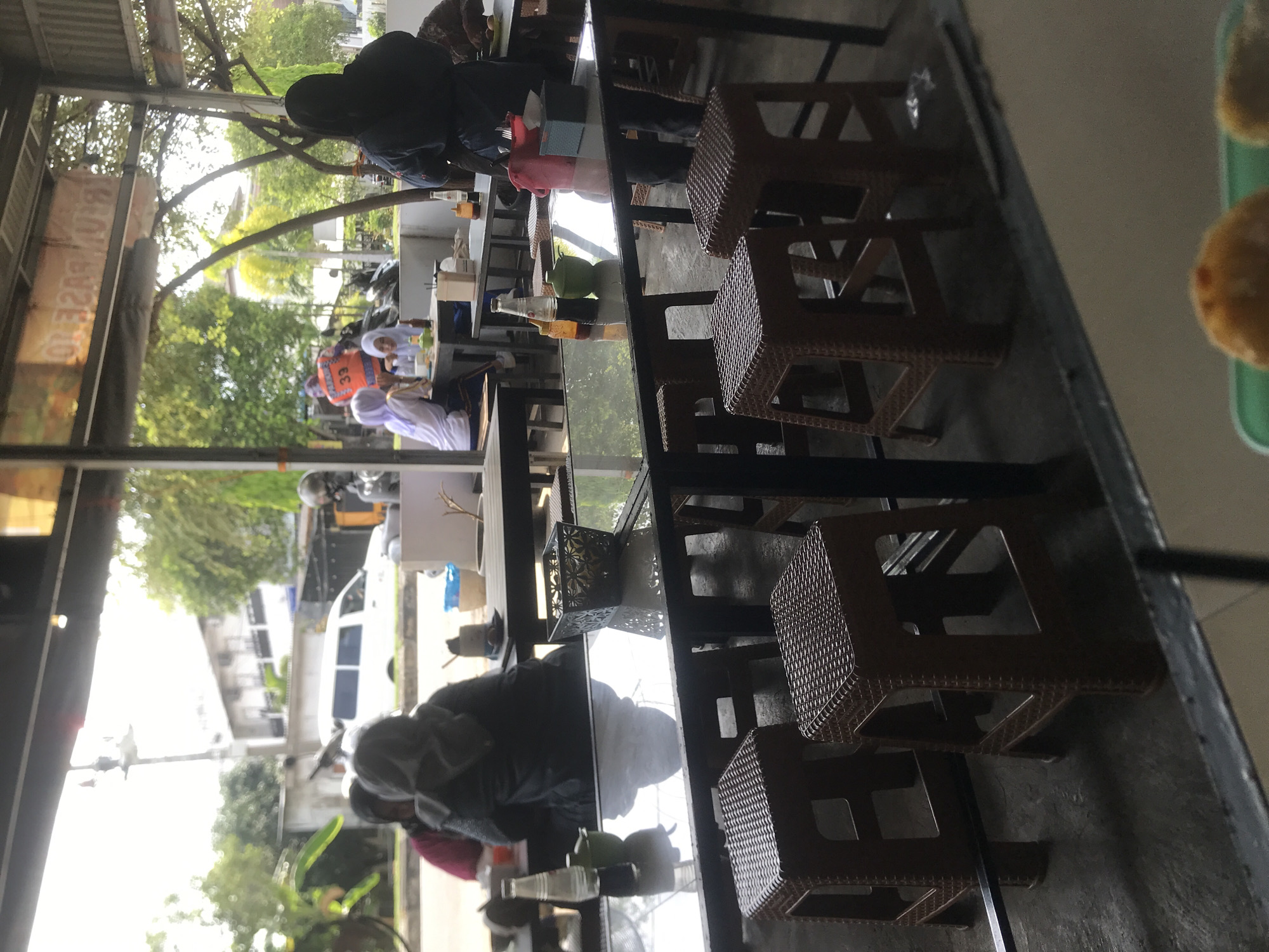 The image shows a view from a restaurant or cafe in Lhokseumawe, Indonesia. Several people are sitting at tables, some wearing headscarves. A white van is visible on the street outside, and there are trees and buildings. Inside the cafe, there are tables with multiple brown rattan stools, a decorative candle holder, and bottles of condiments. A green tray with food is in the foreground.