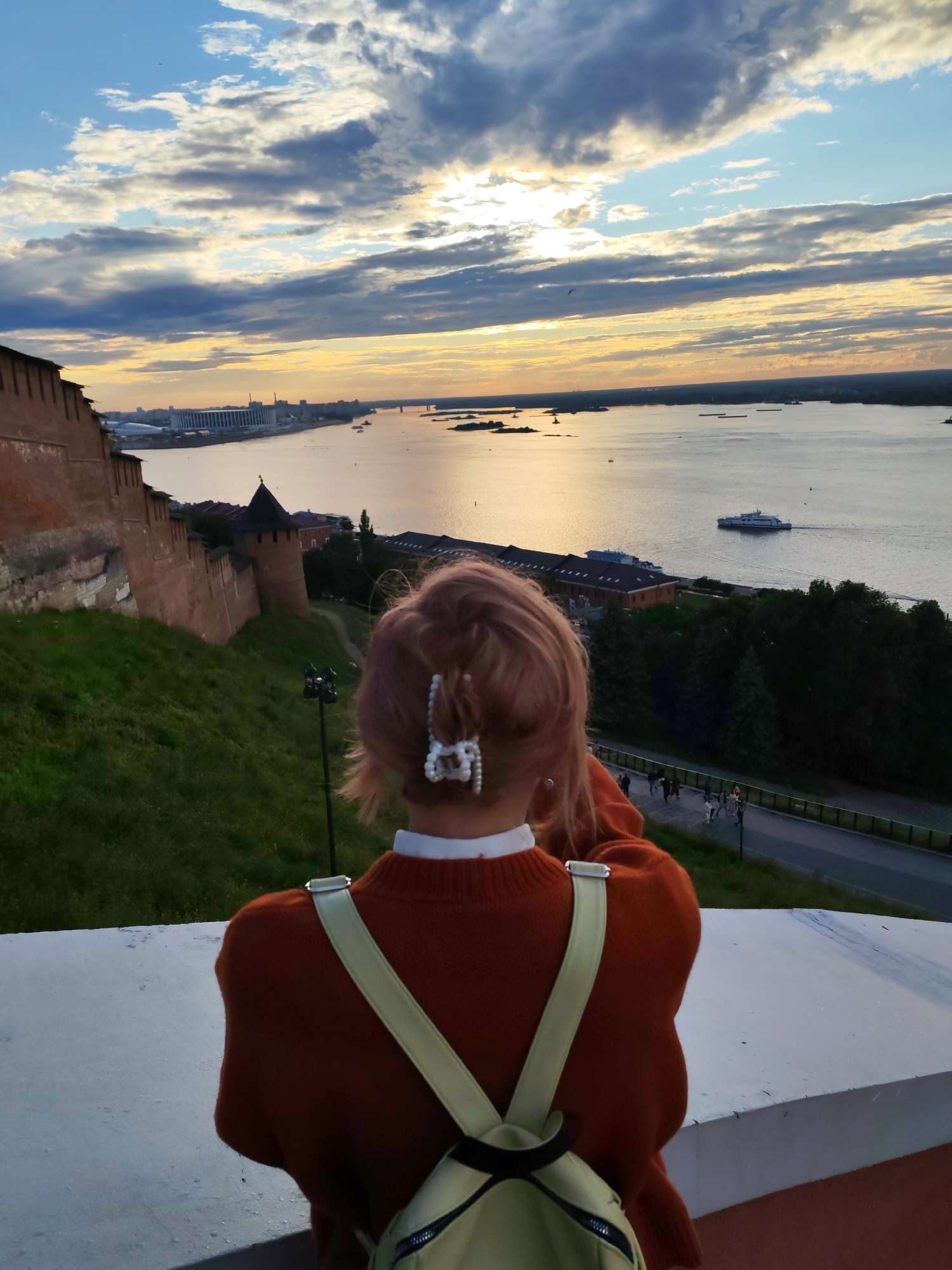 A person with light pink hair, adorned with a pearl hair clip, stands with their back to the camera, facing a panoramic view of the confluence of the Oka and Volga rivers in Nizhny Novgorod, Russia. The sky is filled with dramatic clouds, illuminated by the setting sun, casting a warm glow over the water. In the foreground, the ancient red brick walls of the Nizhny Novgorod Kremlin rise from a grassy slope. The river is dotted with several boats, including a ferry sailing in the distance. Below the Kremlin walls, a pedestrian walkway is visible, with a few people strolling along it. The person in the foreground wears an orange sweater and a cream-colored backpack. The scene captures a serene moment overlooking the historic cityscape and the expansive waterways.