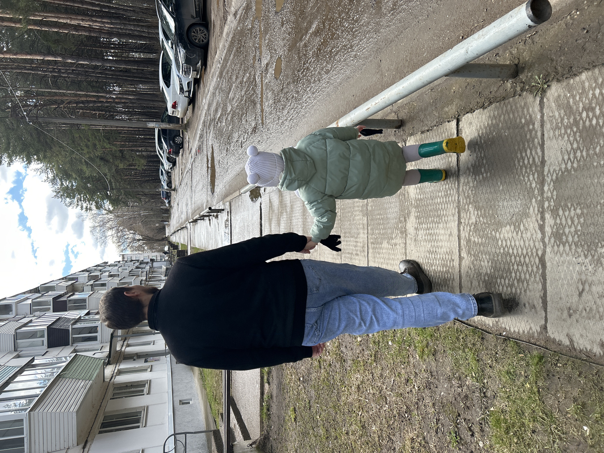 The image shows an adult and a child walking on a paved sidewalk in Chaikovsky, Russia. A multi-story apartment building is visible in the background to the left, and a line of parked cars with a forest behind them runs along the right. The sidewalk is bordered by a metal railing. The adult, seen from the back, wears a black jacket and blue jeans. The child wears a light green jacket, a white hat with small ears, and green and yellow boots. They are holding hands. The weather appears to be wet or muddy.