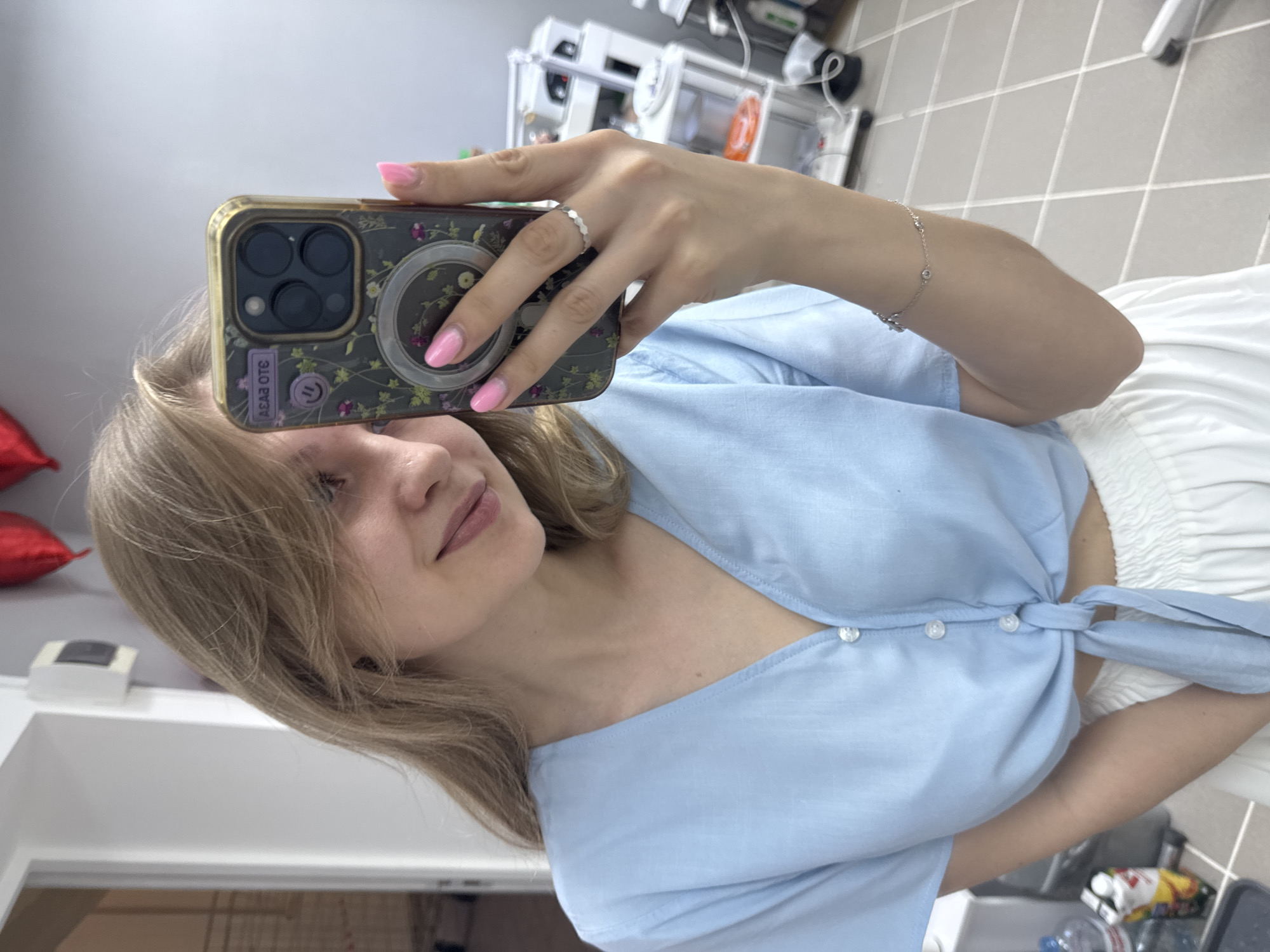 A young woman with blonde hair and light skin is taking a selfie with her iPhone. She is wearing a light blue button-down shirt tied at the front and a white skirt. Her nails are painted pink, and she wears a silver ring and bracelet. The phone has a patterned case with three camera lenses and a magnetic ring. The background shows a room with grey tiled floors, medical equipment, and a window. Red balloons are visible in the corner. The location is potentially in Izhevsk, Russia.