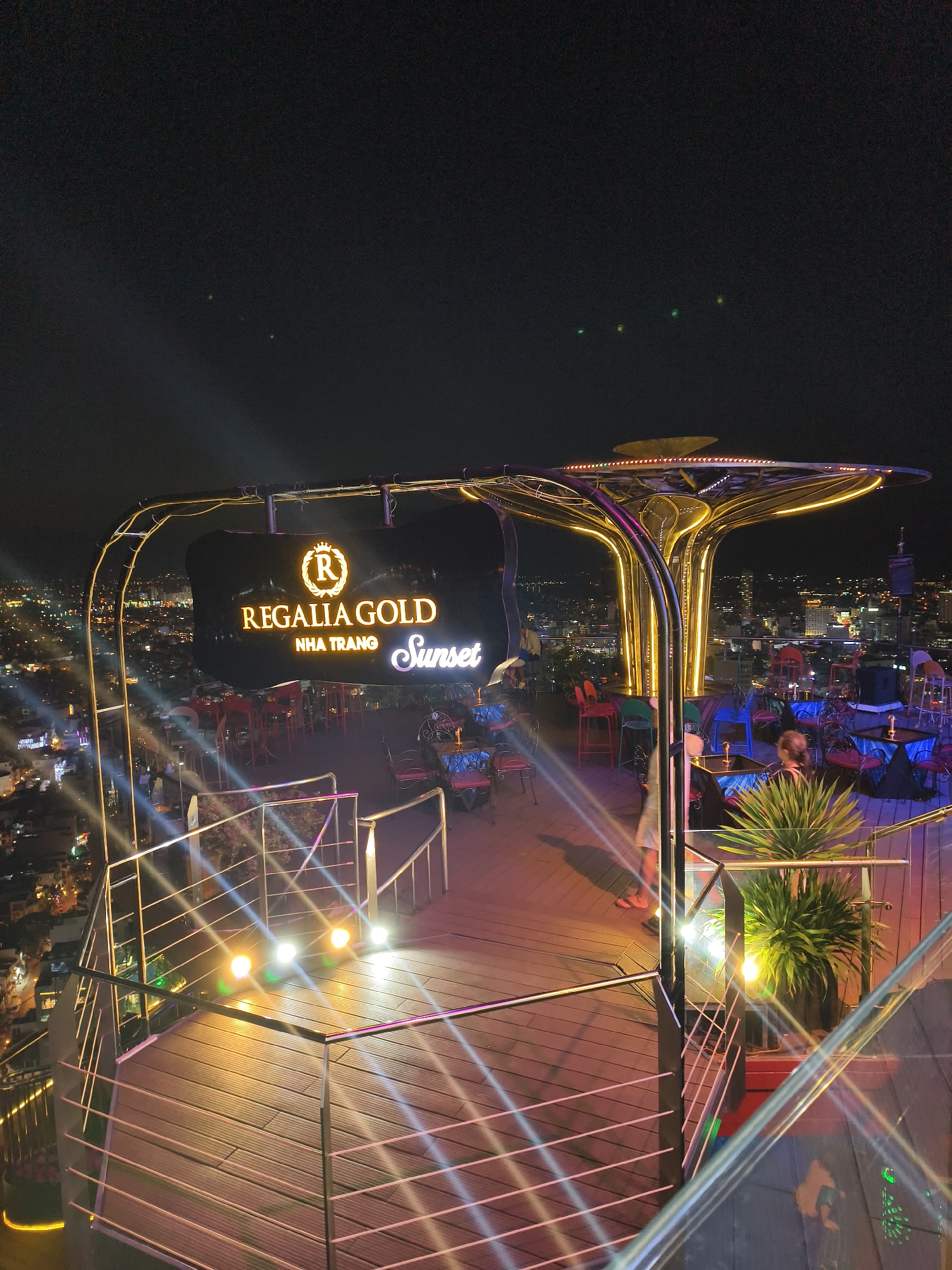 This image captures a vibrant rooftop bar or restaurant at night, offering panoramic views of a sprawling city illuminated by countless lights. The time of day is clearly evening, with a dark, clear sky above. The setting is urban, with the city lights extending to the horizon, suggesting a high vantage point from a tall building.
The central subject is the rooftop establishment itself, featuring a large, brightly lit sign with glowing text that reads "REGALIA GOLD" at the top, "NHA TRANG" below it, and "Sunset" in a stylized script to the right. This text directly indicates the venue's name and its location: Nha Trang.
The bar area is furnished with numerous small tables and chairs in various colors, including red, blue, and green, scattered across a wooden deck. Several people are visible, some seated at tables appearing to socialize, while others are partially seen moving around. One person is visible near a large potted plant in the foreground, and another figure in a light-colored shirt is seen walking on the deck.
A distinctive architectural feature is a large, abstract, tree-like structure with a metallic, golden-colored finish and integrated lighting that glows warmly, creating a unique focal point. The deck is lined with modern metal railings and glass panels, allowing for unobstructed views of the city. Additional floor spotlights cast bright, multi-colored light patterns across the wooden surface and railings. Potted plants are strategically placed, adding touches of greenery to the otherwise man-made environment. The overall atmosphere appears lively, modern, and inviting.